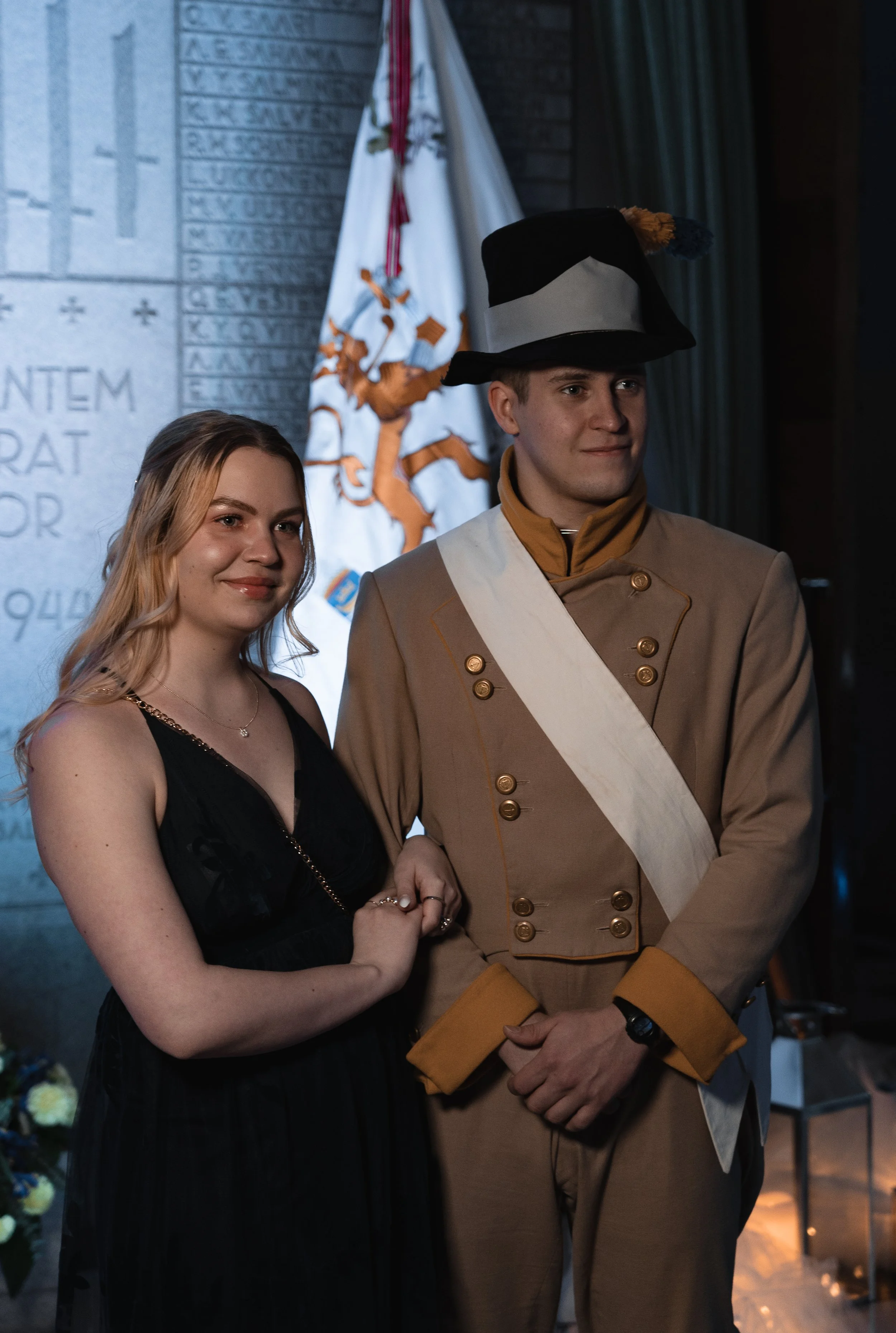 A young woman in a black dress standing next to a young man in historical military uniform, holding hands at a formal event with a flag and a stone wall in the background.
