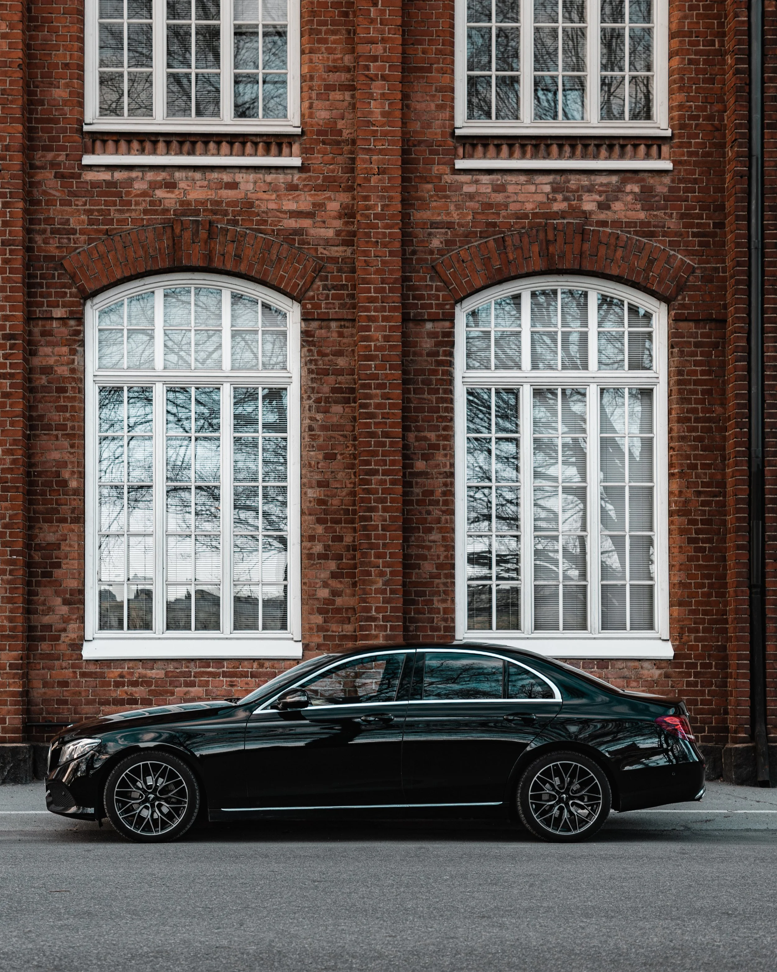 A sleek black sedan parked on a street in front of a red brick building with large, white-framed multi-pane windows.