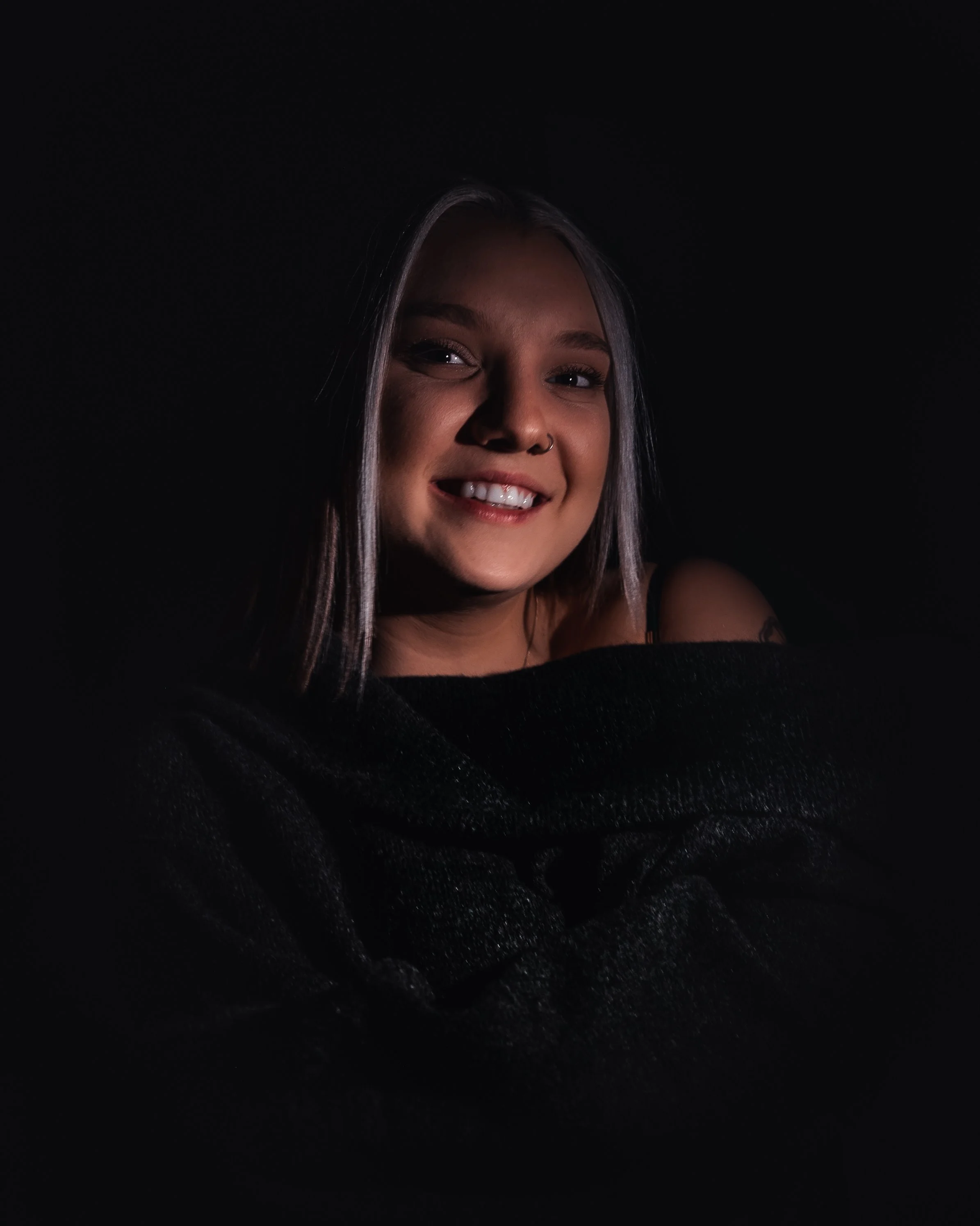 A young woman with gray hair and a nose ring, smiling, wearing a black off-the-shoulder top, against a dark background.