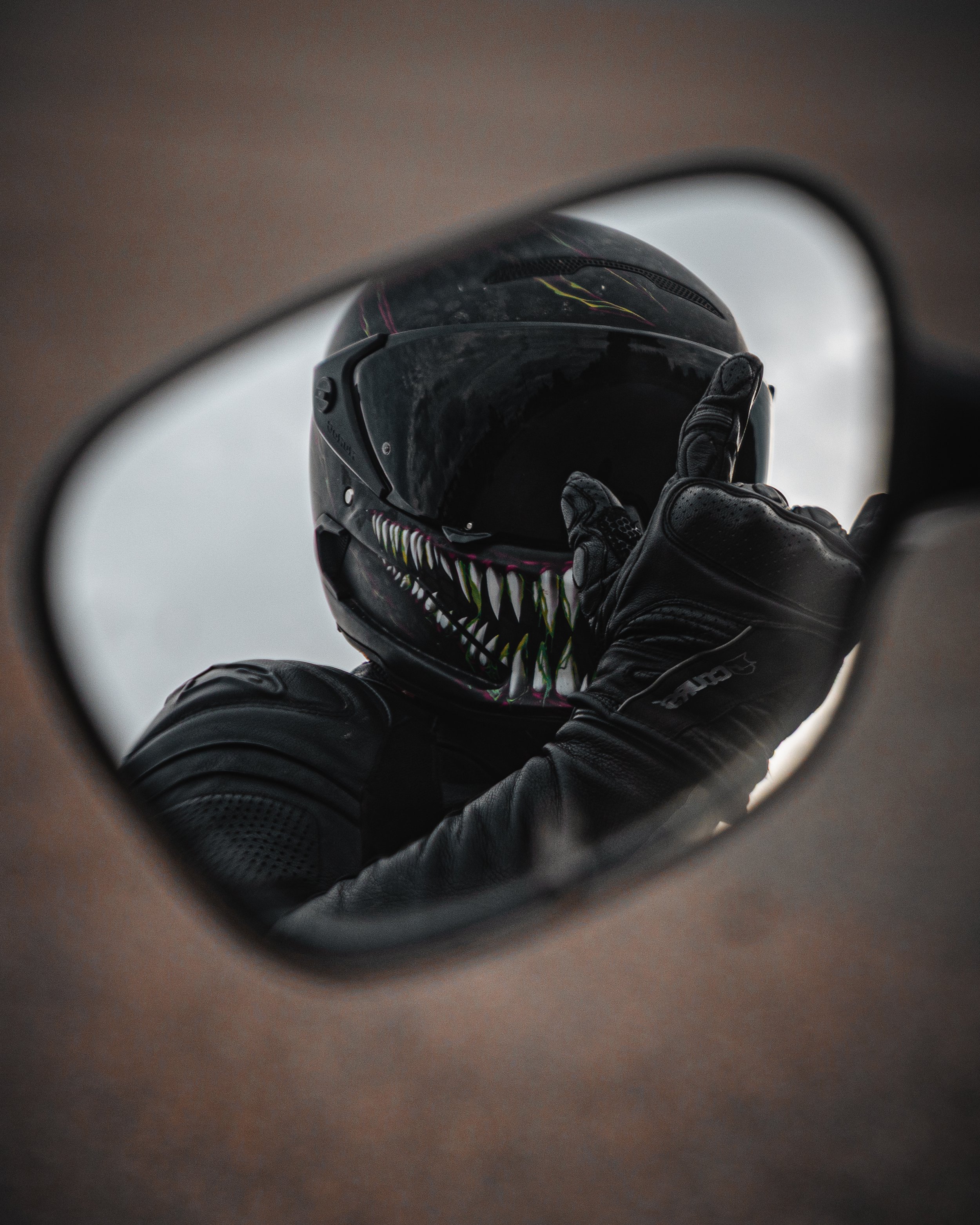 A person wearing a black motorcycle helmet with colorful teeth design and black gloves, showing an 'okay' hand gesture through a mirror.