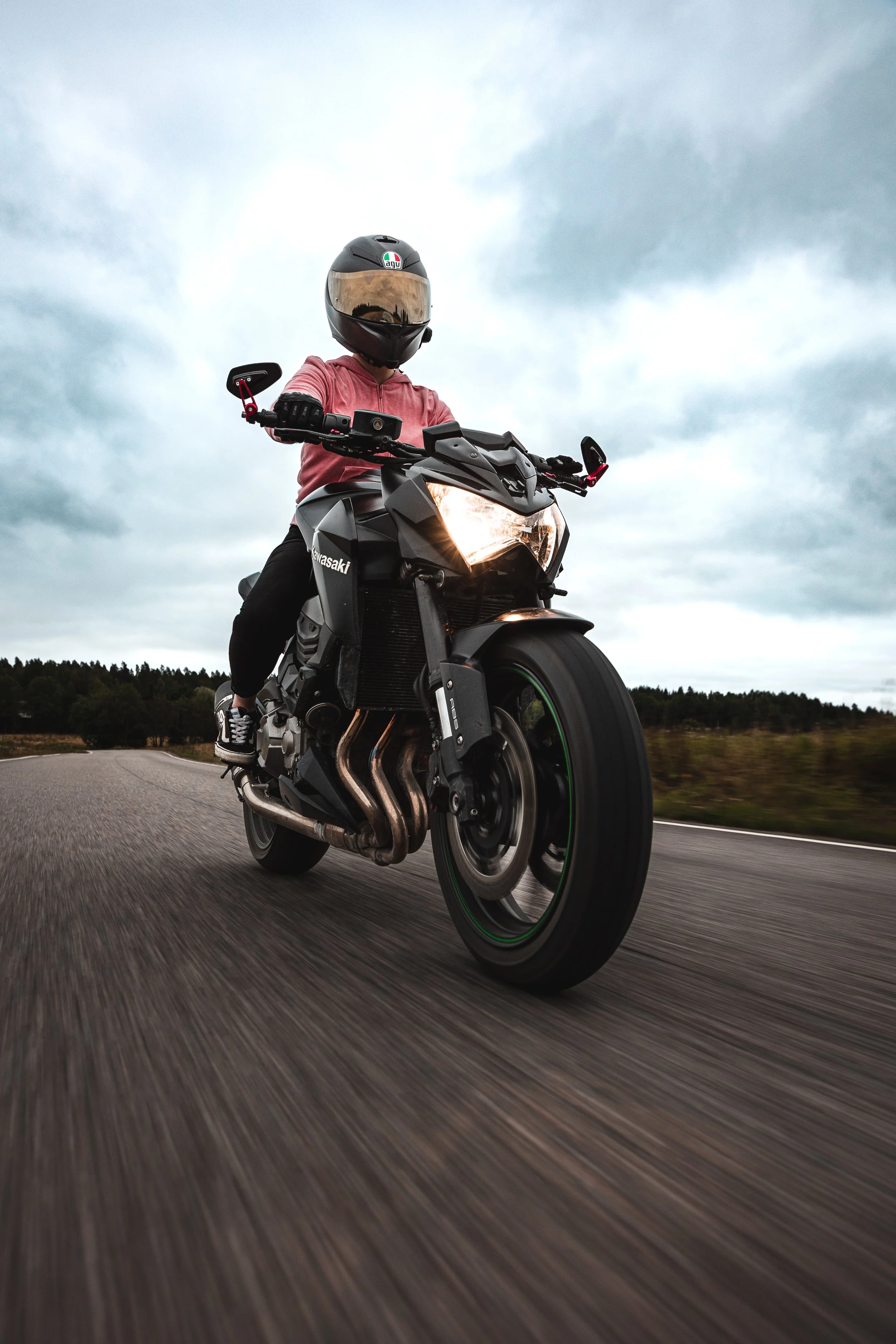 A person wearing a helmet and pink hoodie riding a black Kawasaki motorcycle on an open road under cloudy sky.