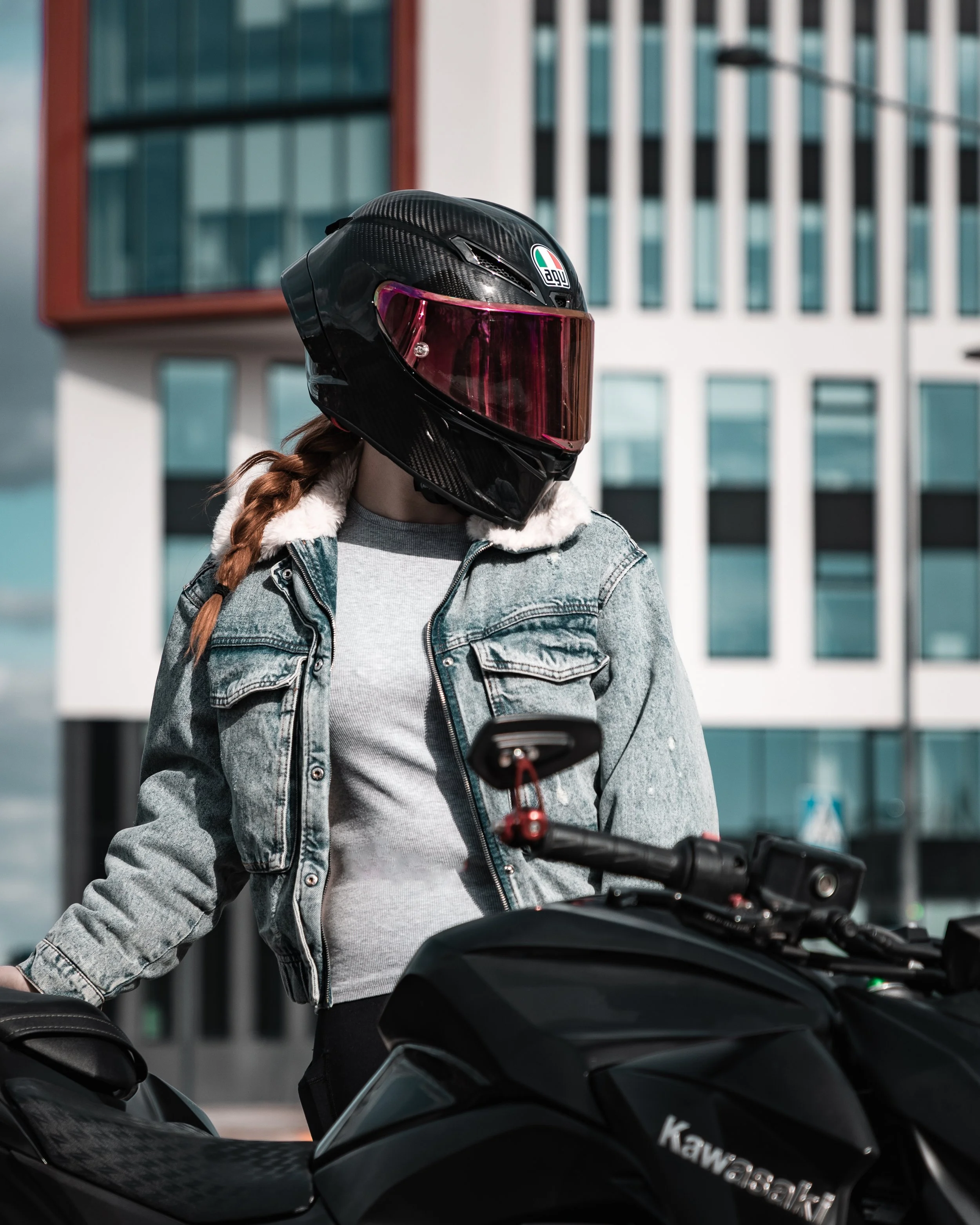 A woman wearing a black motorcycle helmet with a pink visor, a gray hoodie, and a denim jacket standing next to a black Kawasaki motorcycle in an urban setting.