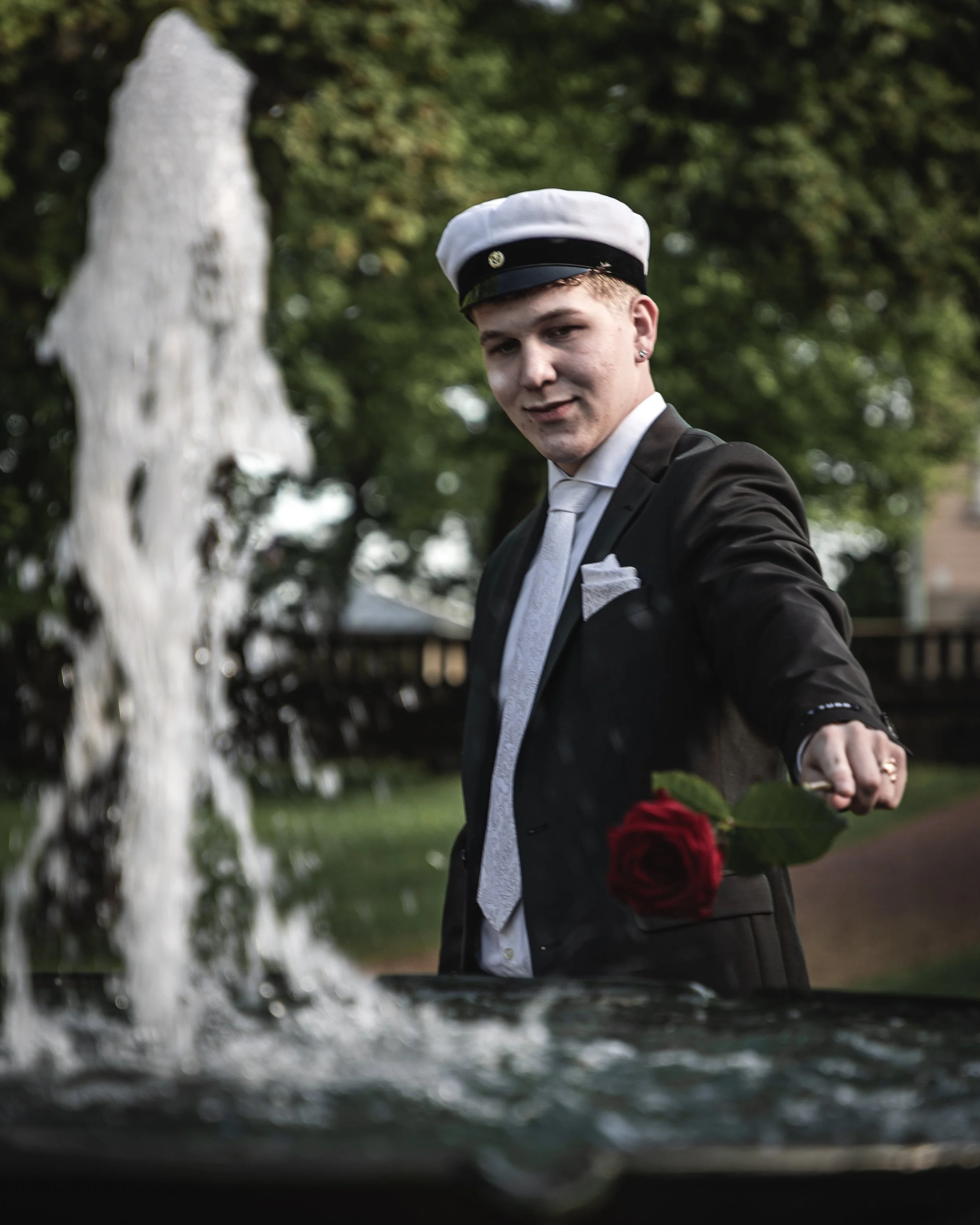 Young person in formal attire with a white cap, holding a red rose, standing outdoors near a fountain with water splashing, and trees in the background.