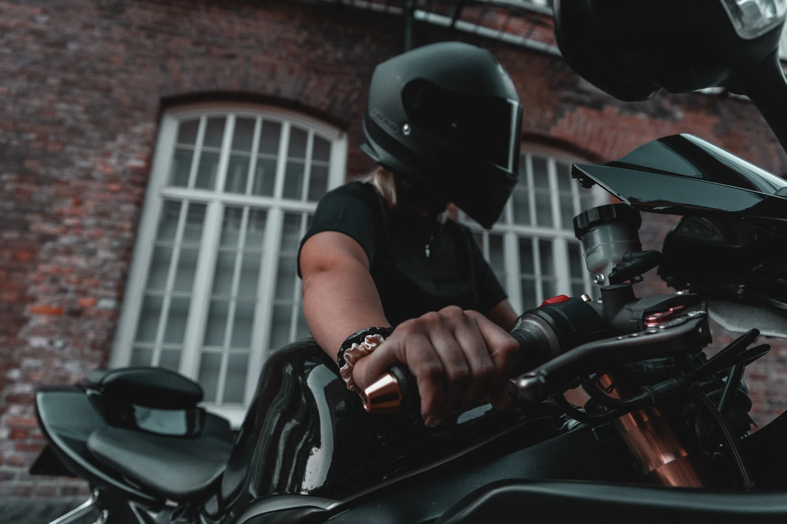 Person wearing a black motorcycle helmet and black shirt, sitting on a black motorcycle, with a brick building with white window frames in the background.