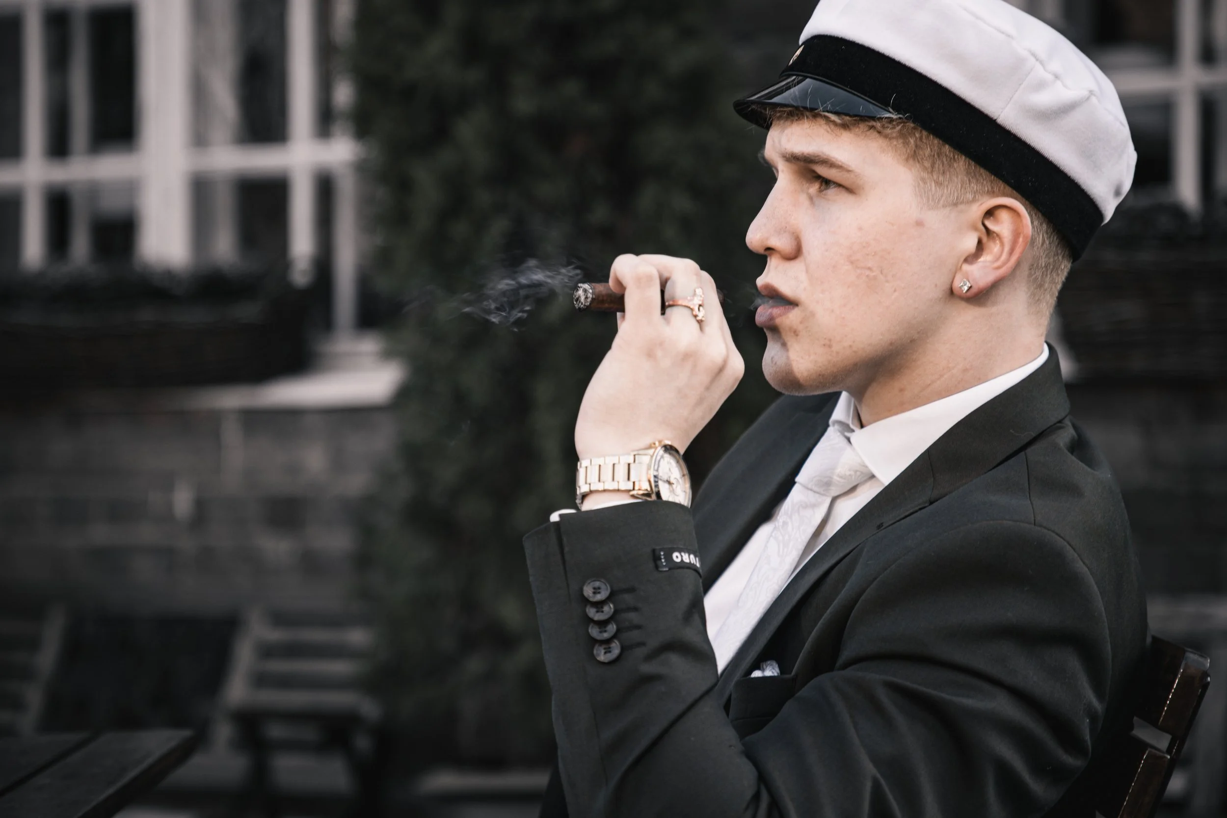 Young man in a suit, white shirt, and a sailor cap, smoking a cigar outside with a blurred background.