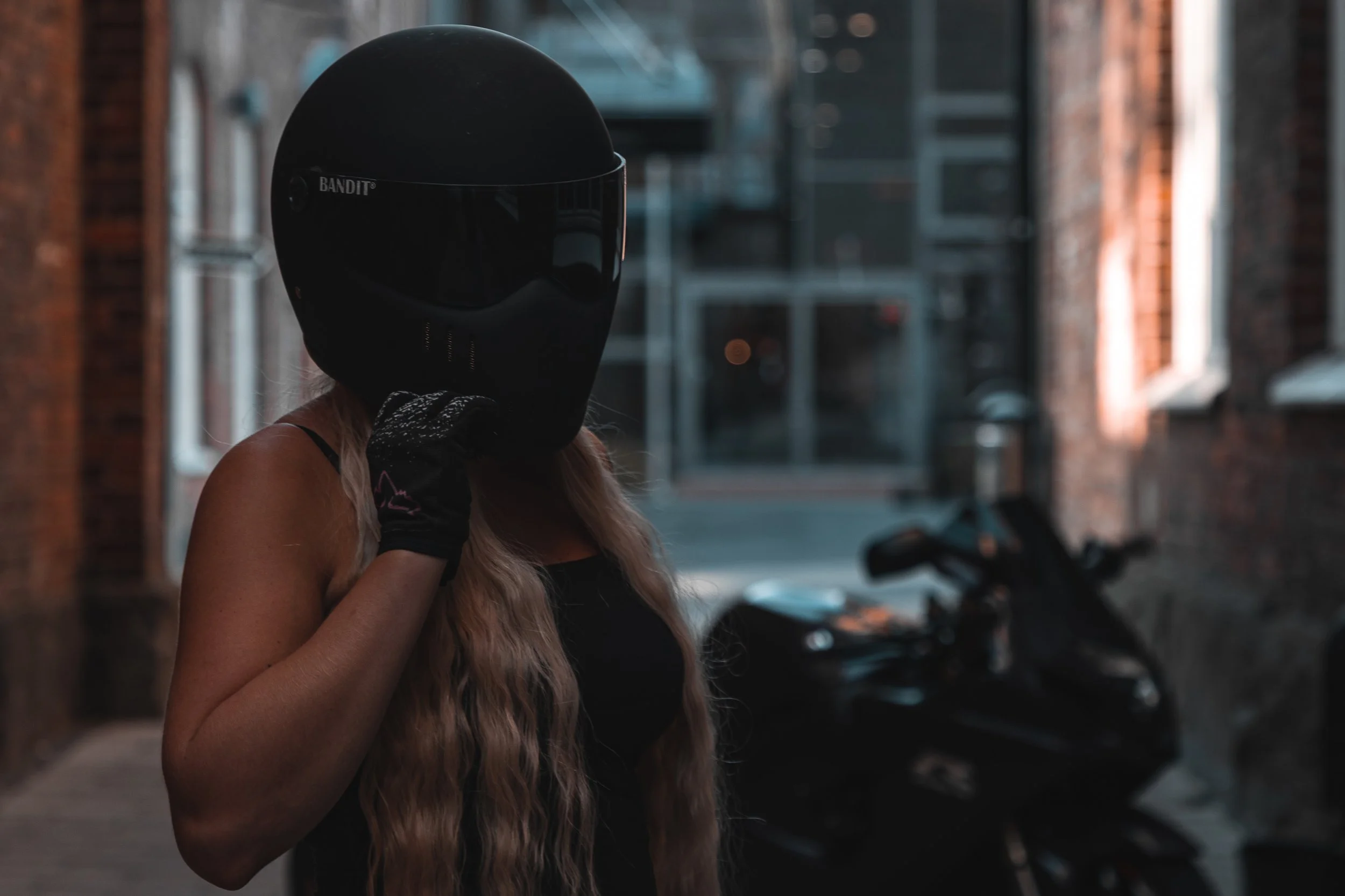 A woman wearing a black motorcycle helmet and black gloves, standing next to a motorcycle in an alleyway with brick walls.