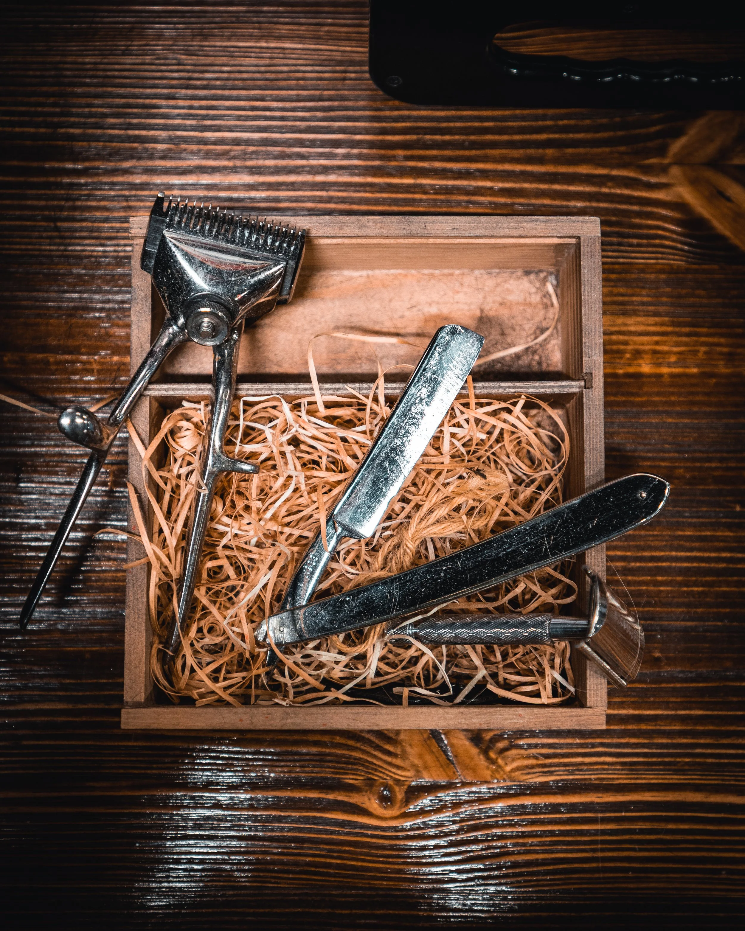 A set of barber tools including scissors, a straight razor, and a clipper, placed in a wooden box with wood shavings on a wooden surface.