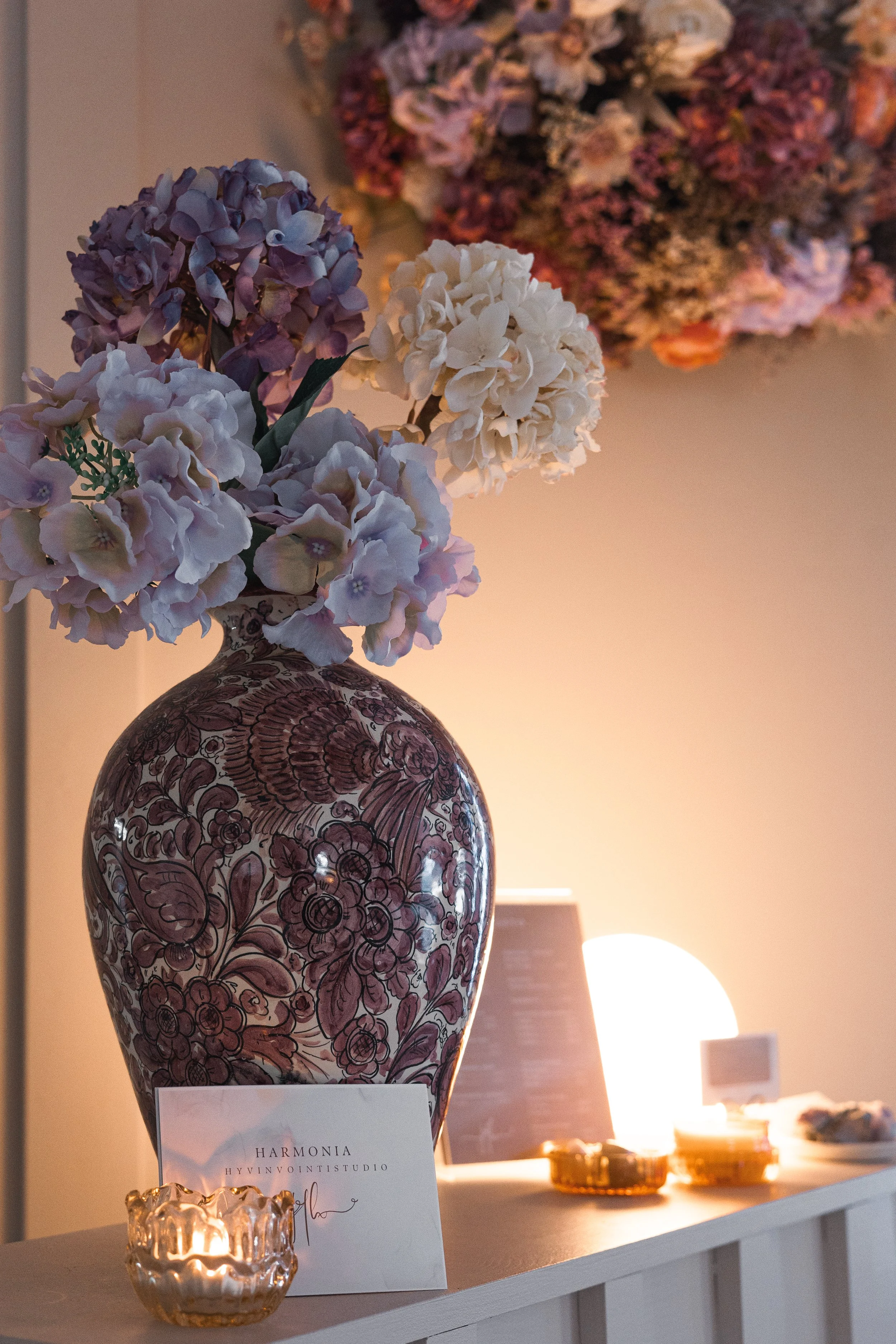Vase with purple and white hydrangea flowers on a table with a small candle holder, a card with text, and a lamp in the background, with a large floral arrangement on the wall behind.
