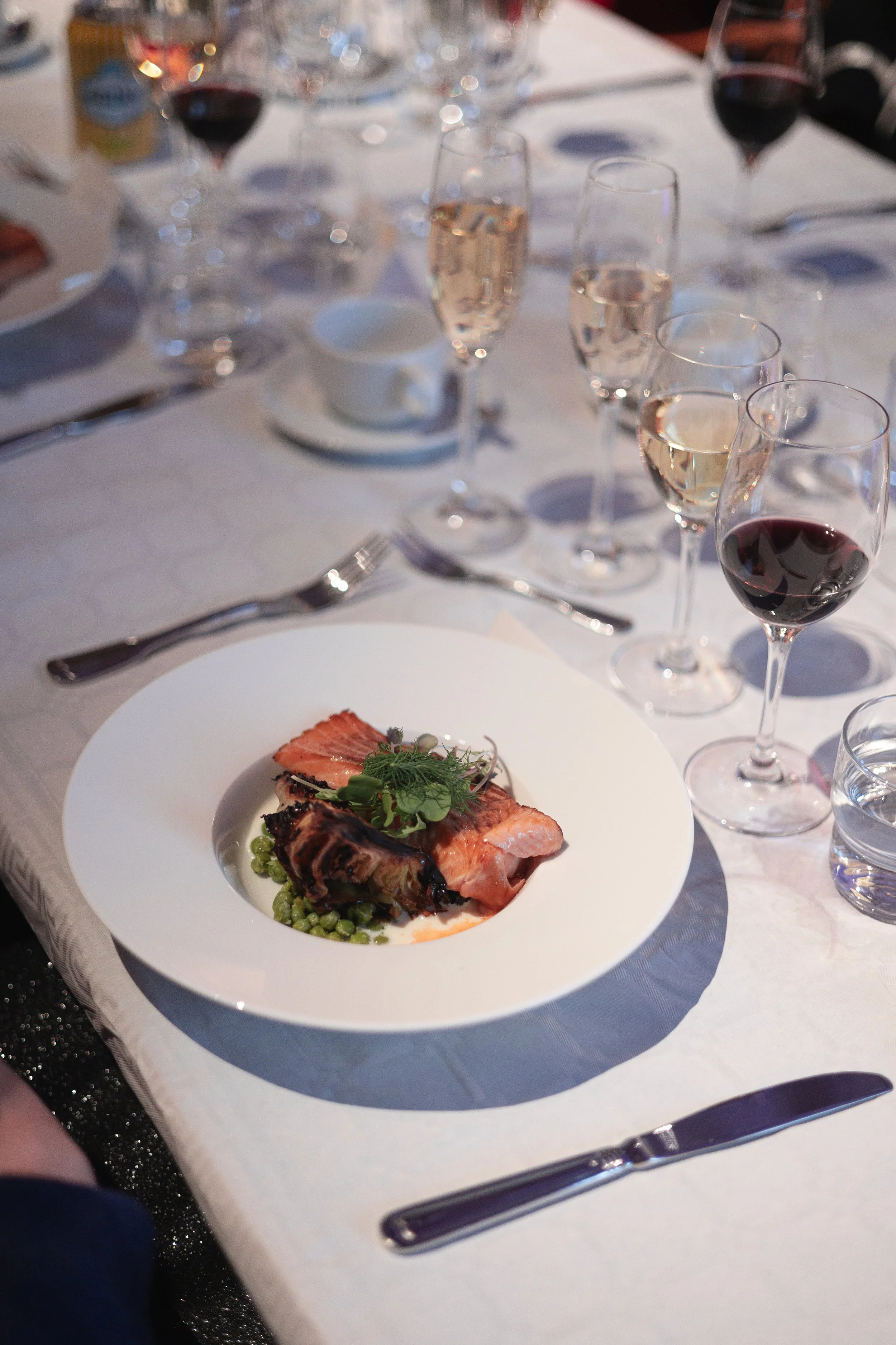 A dinner table set with wine glasses, champagne flutes, silverware, and a white plate with a piece of cooked salmon garnished with greens, served on a white tablecloth.