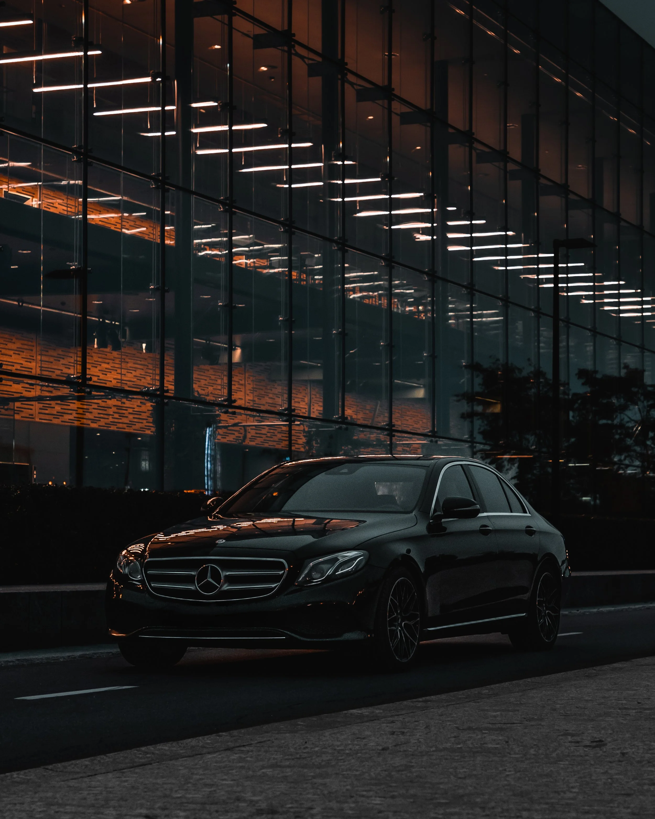 A black Mercedes-Benz sedan parked on the street at night in front of a modern glass building with illuminated interior lighting.