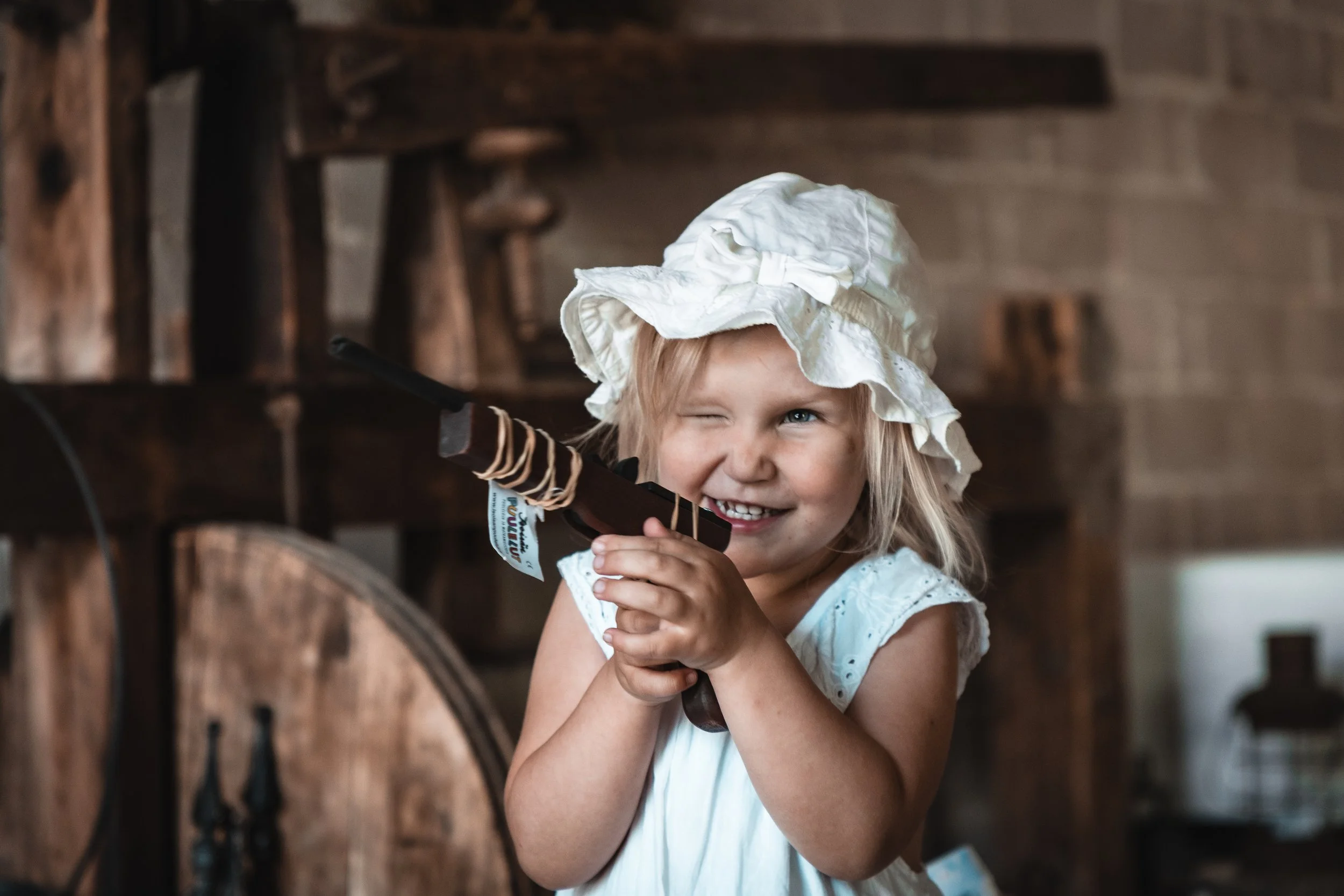 A young girl wearing a white bonnet and dress, smiling and holding a toy rifle indoors.