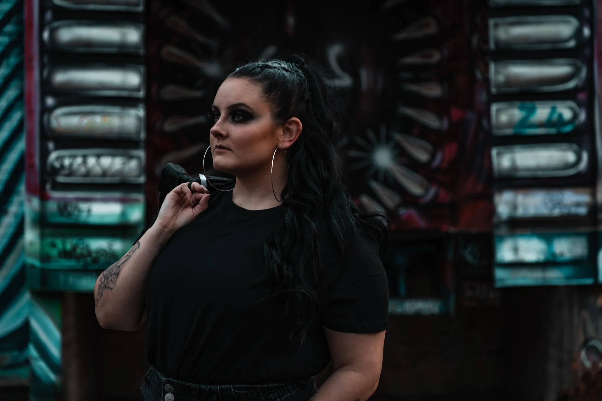 A woman with dark hair, wearing a black shirt and large hoop earrings, holding a smartphone to her shoulder while standing in front of a graffiti-covered wall.