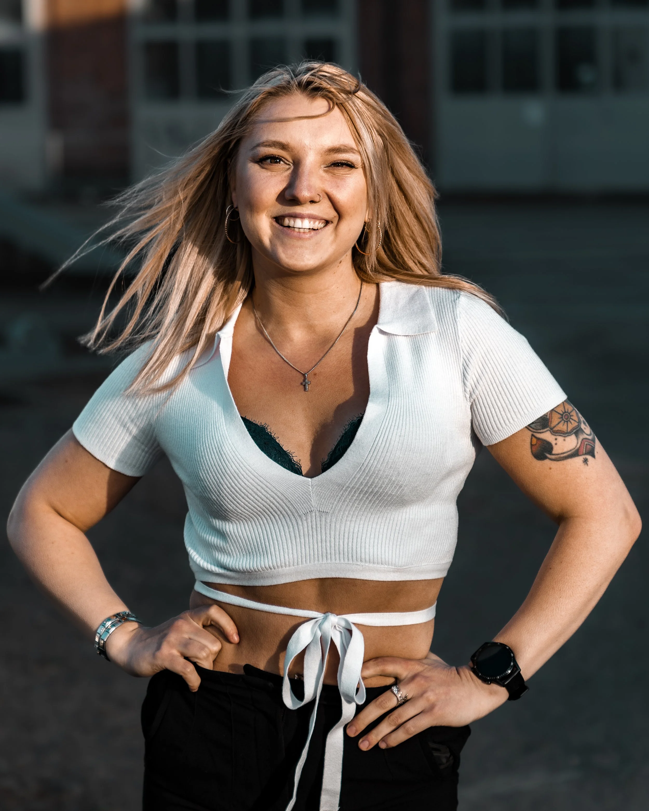 A smiling young woman with blonde hair, wearing a white crop top tied at the waist, black lace bra underneath, and black pants. She has tattoos on her right arm, a necklace with a cross, and a smartwatch on her left wrist. She is standing outdoors wi