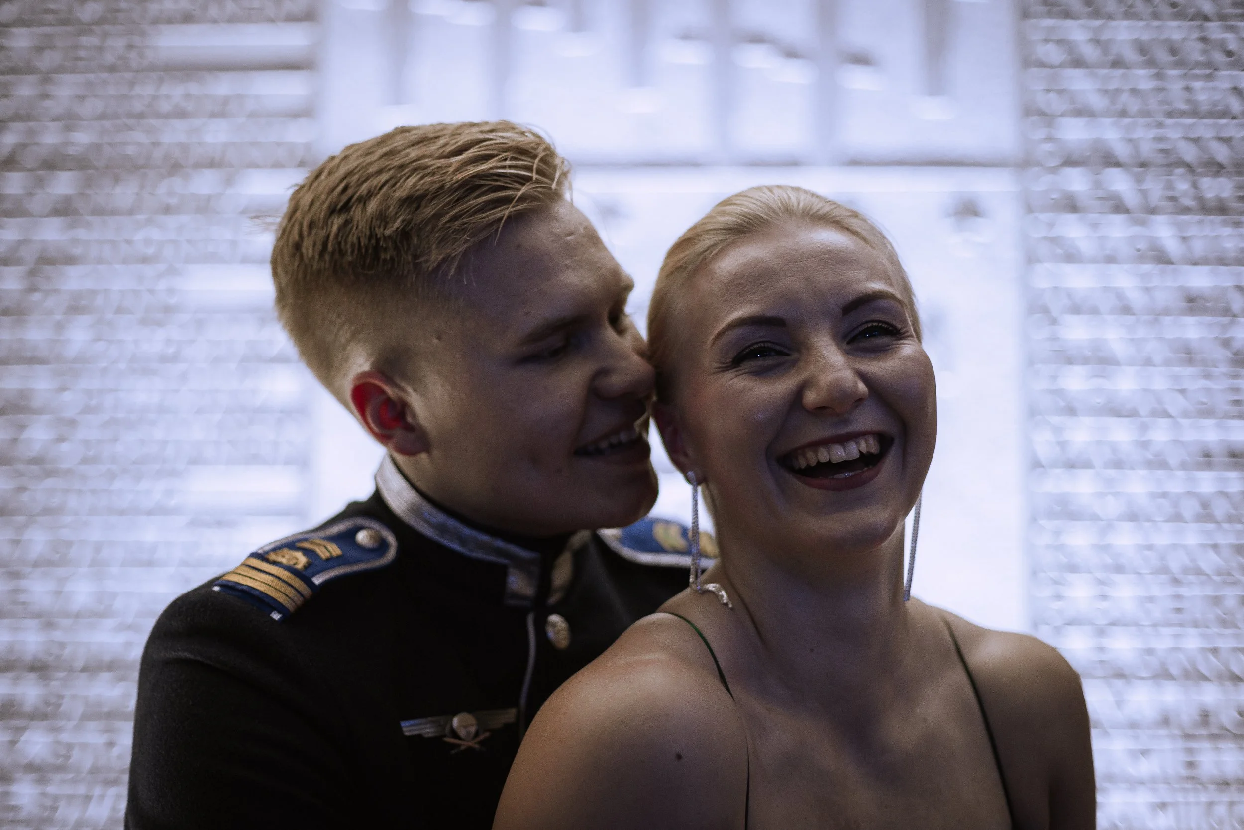 A man in a military uniform whispers into a woman's ear, both smiling and laughing joyfully.