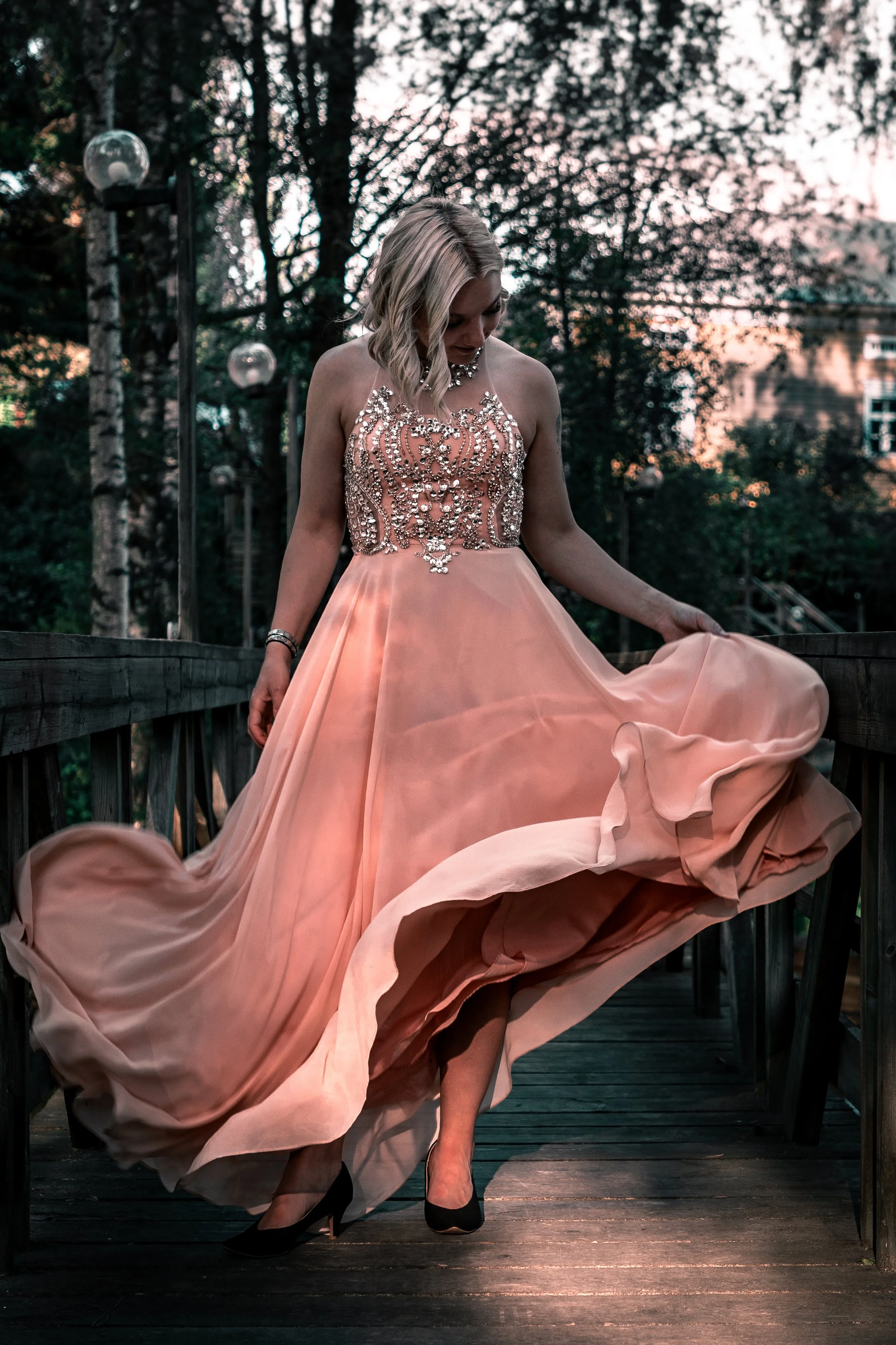 A woman in a flowing peach-colored gown with intricate embellishments on the bodice, standing on a wooden bridge surrounded by trees at sunset.
