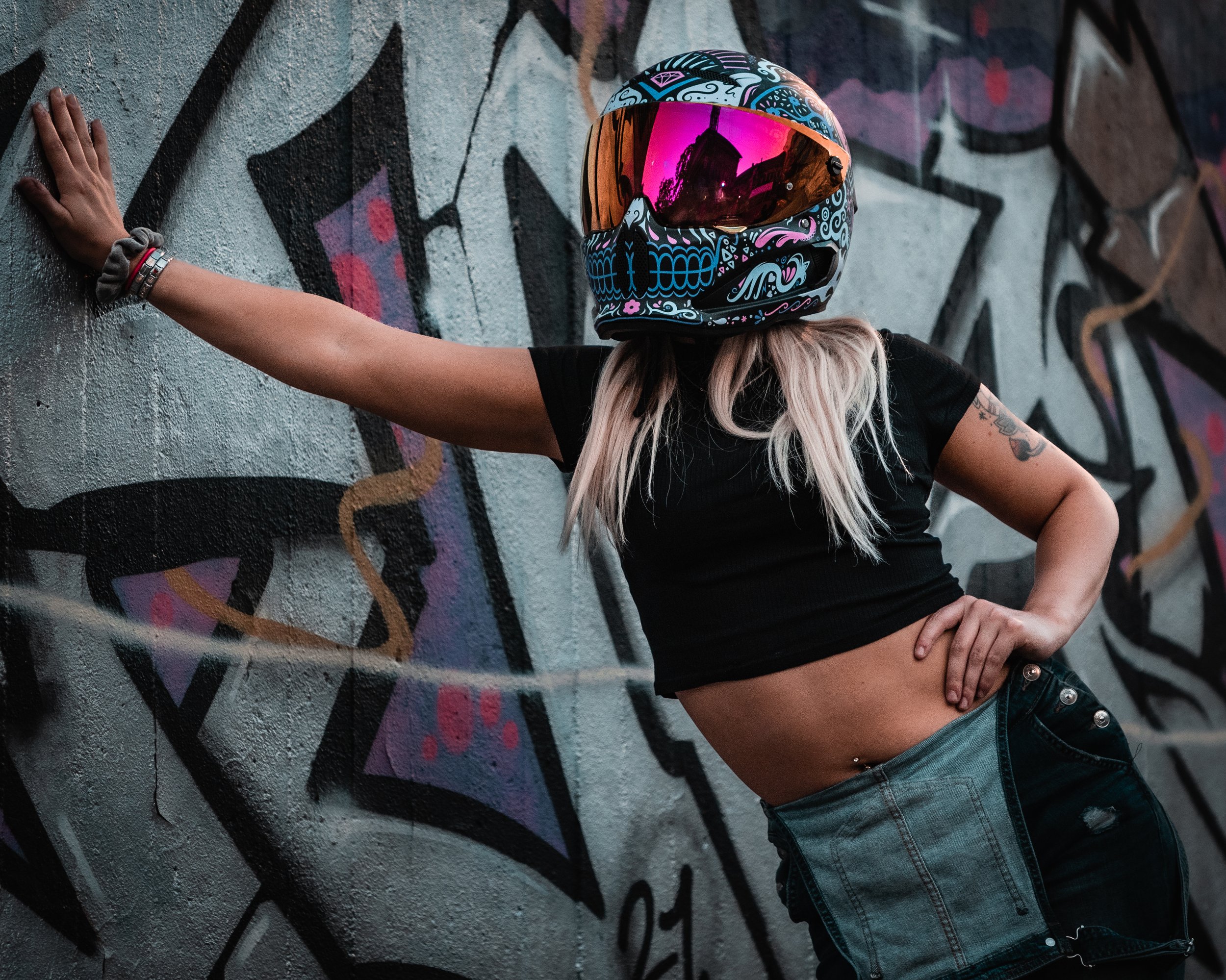 Woman wearing a colorful motorcycle helmet with reflective visor, black crop top, and blue jeans, leaning against a graffiti-covered wall with her hand on her hip.