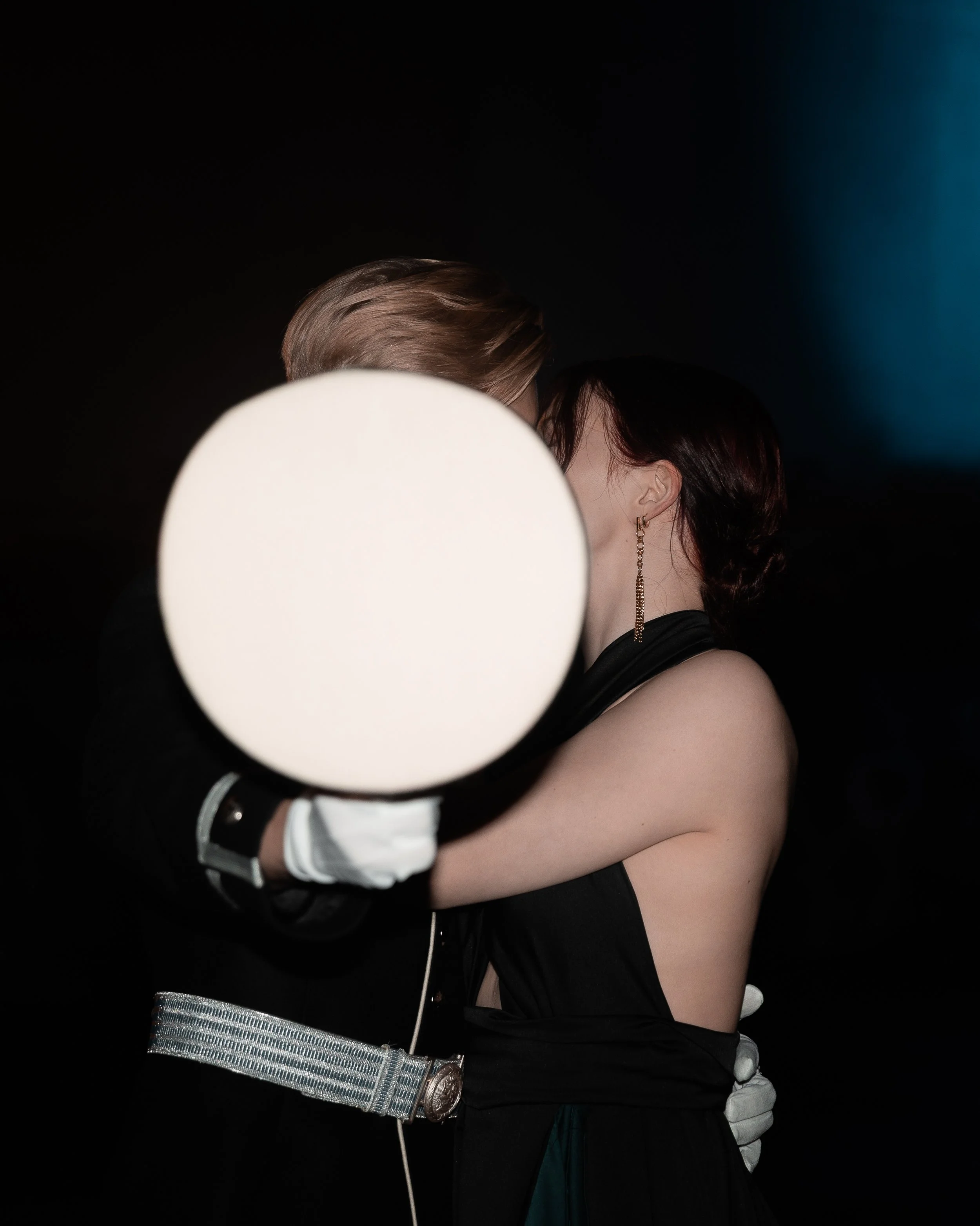A couple kissing, partially obscured by a glowing circular object, with a dark background.