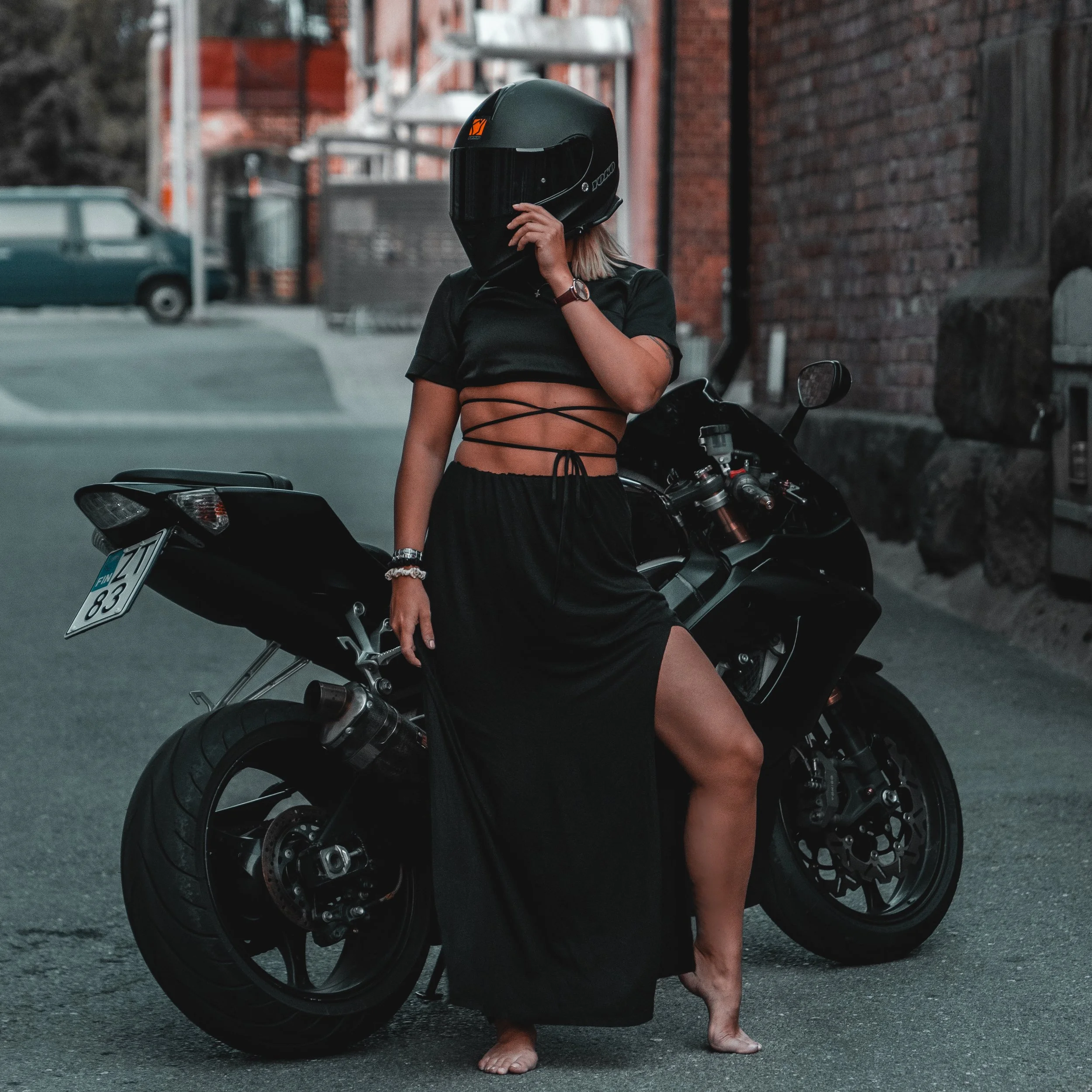 A woman wearing a black motorcycle helmet, black crop top, and long black skirt stands barefoot next to a black sport motorcycle on an urban street with brick buildings.