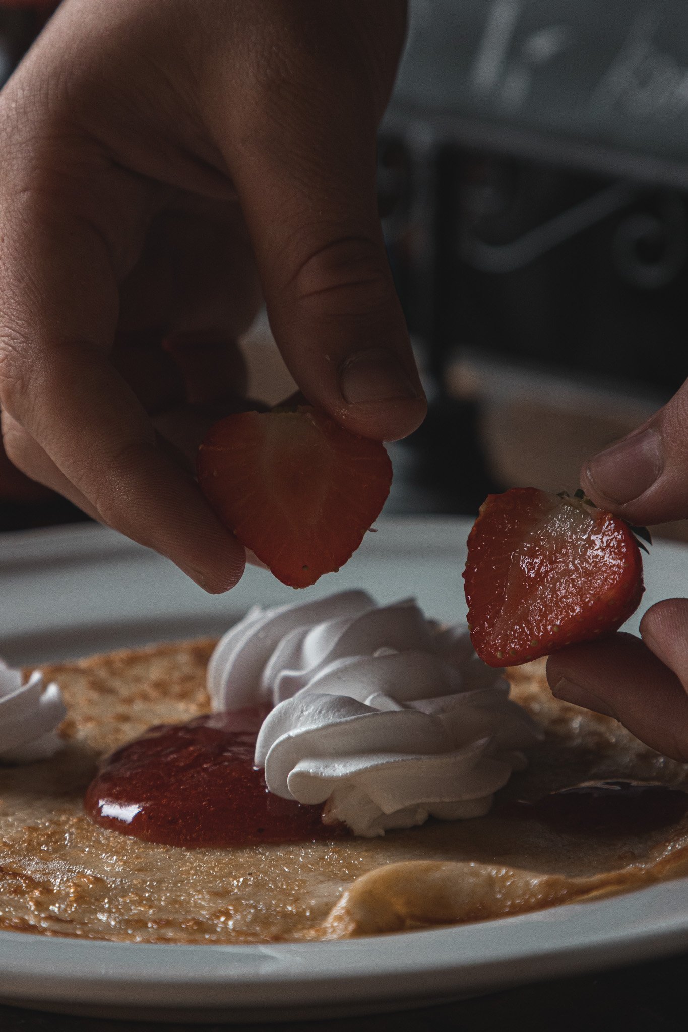 Hands decorating a pancake with strawberry slices, whipped cream, and strawberry syrup.