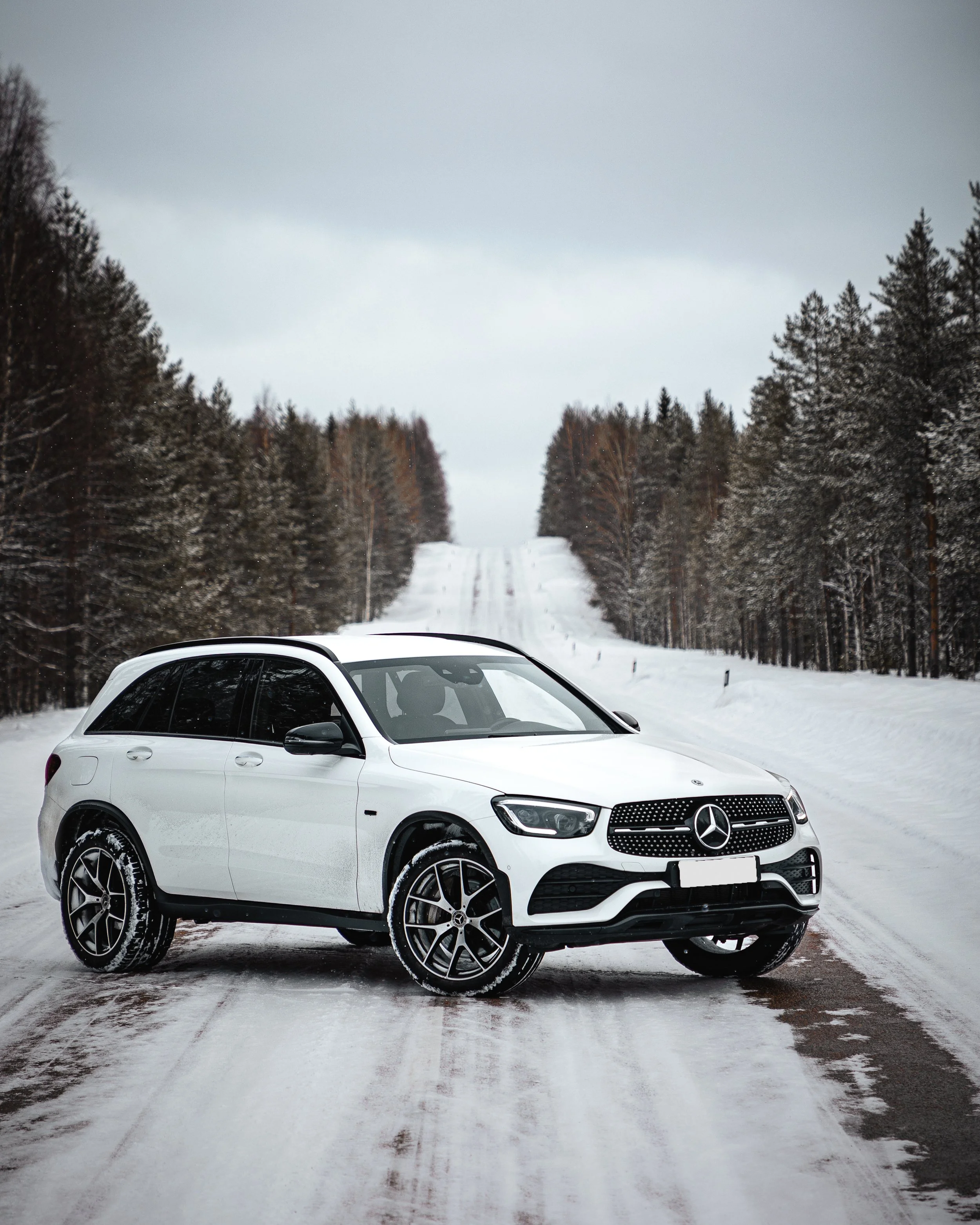 A white Mercedes-Benz SUV parked on a snow-covered road in a forest with leafless trees, under a cloudy sky.