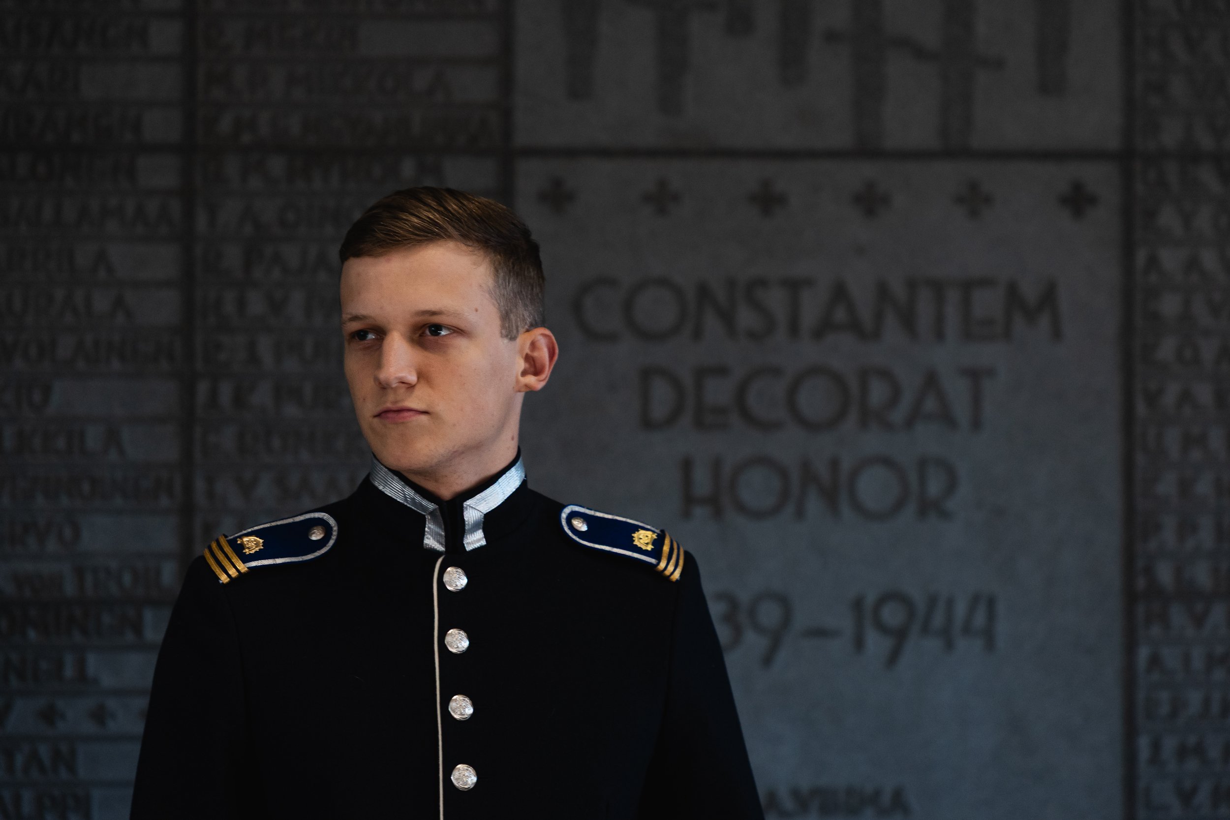 Young man in military uniform standing in front of a memorial wall with the words "Constantem Decorat Honor" and years 1939-1944 engraved on it.