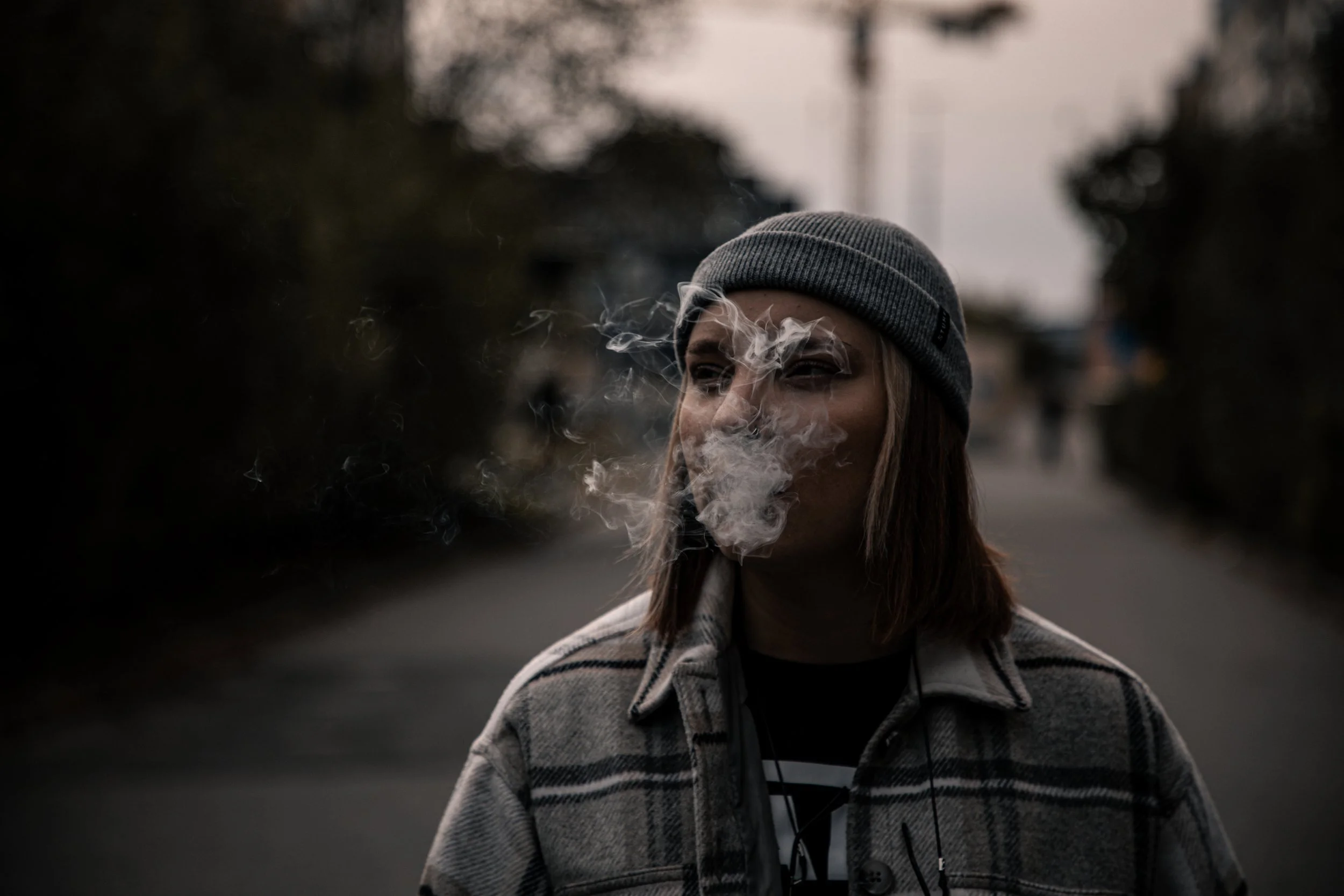A young woman wearing a gray beanie and plaid jacket smokes outdoors during dusk, with smoke partially covering her face and a blurred background of a road and trees.