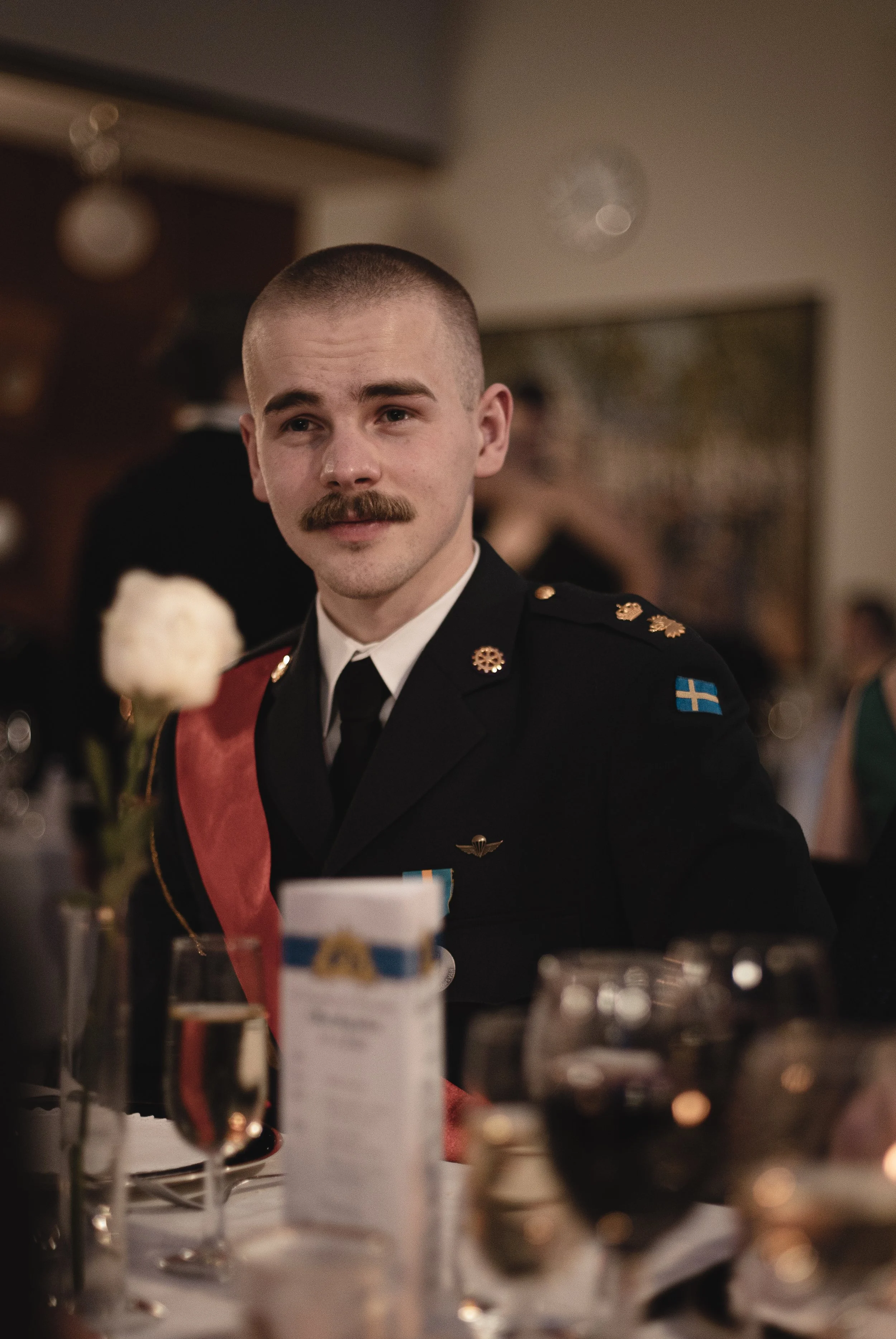 A young man in a military uniform with a mustache, sitting at a table during a formal event, with a blurred background and table setting in the foreground.