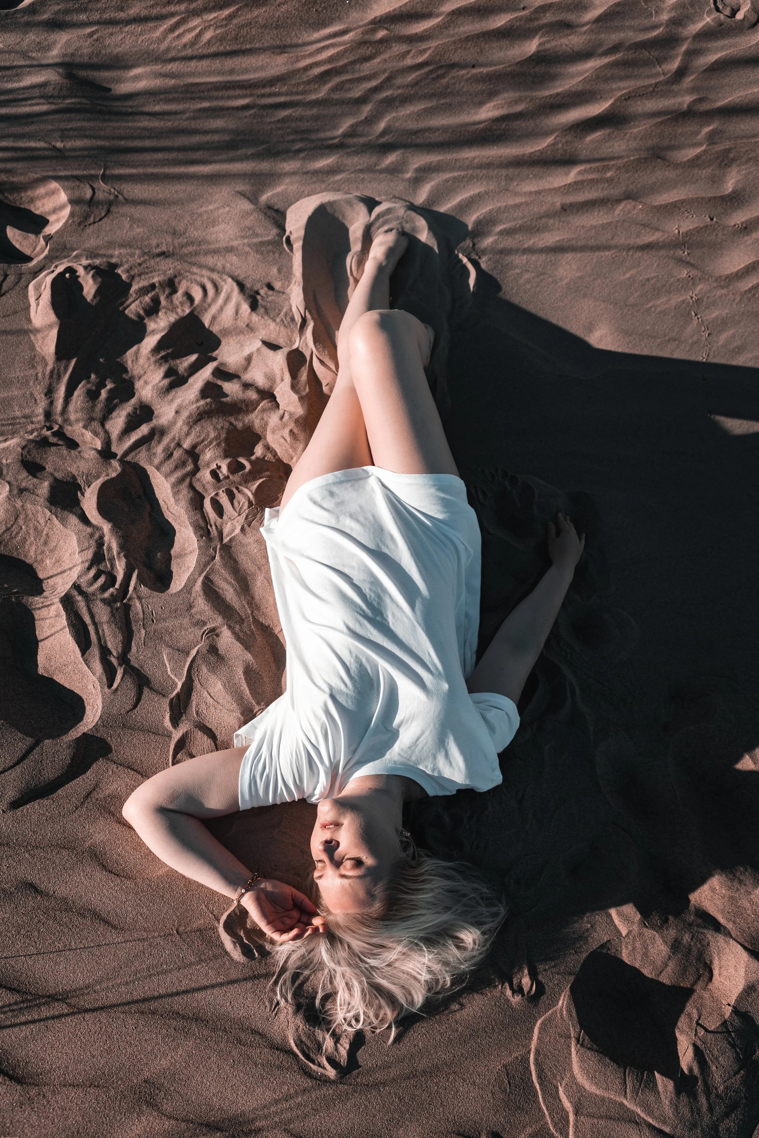 Woman lying on sand in a white dress with her eyes closed, surrounded by shadows and footprints.