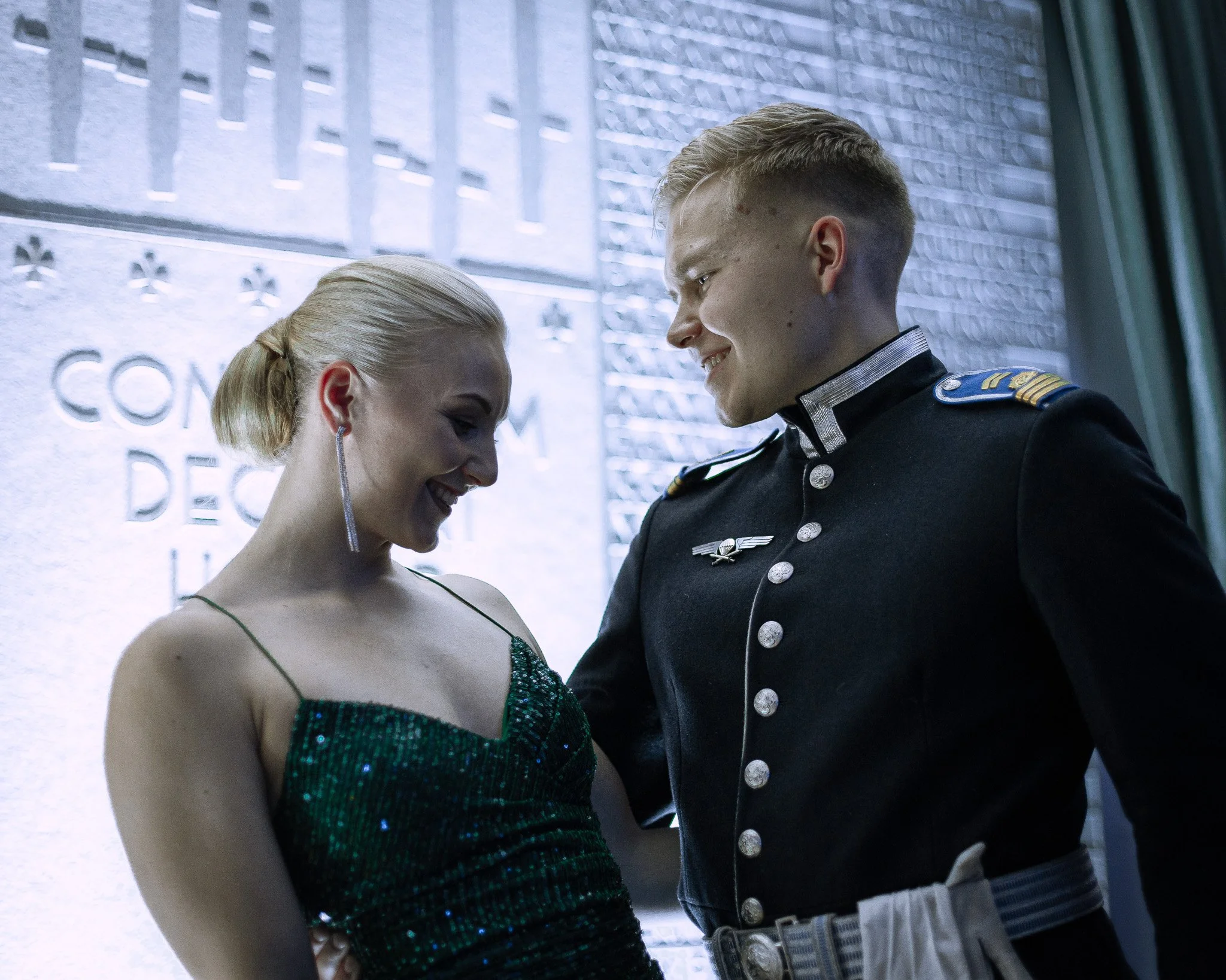 A young woman in a sparkly green dress smiling and looking down, standing close to a young man in a military dress uniform, also smiling, in front of a wall with a plaque.