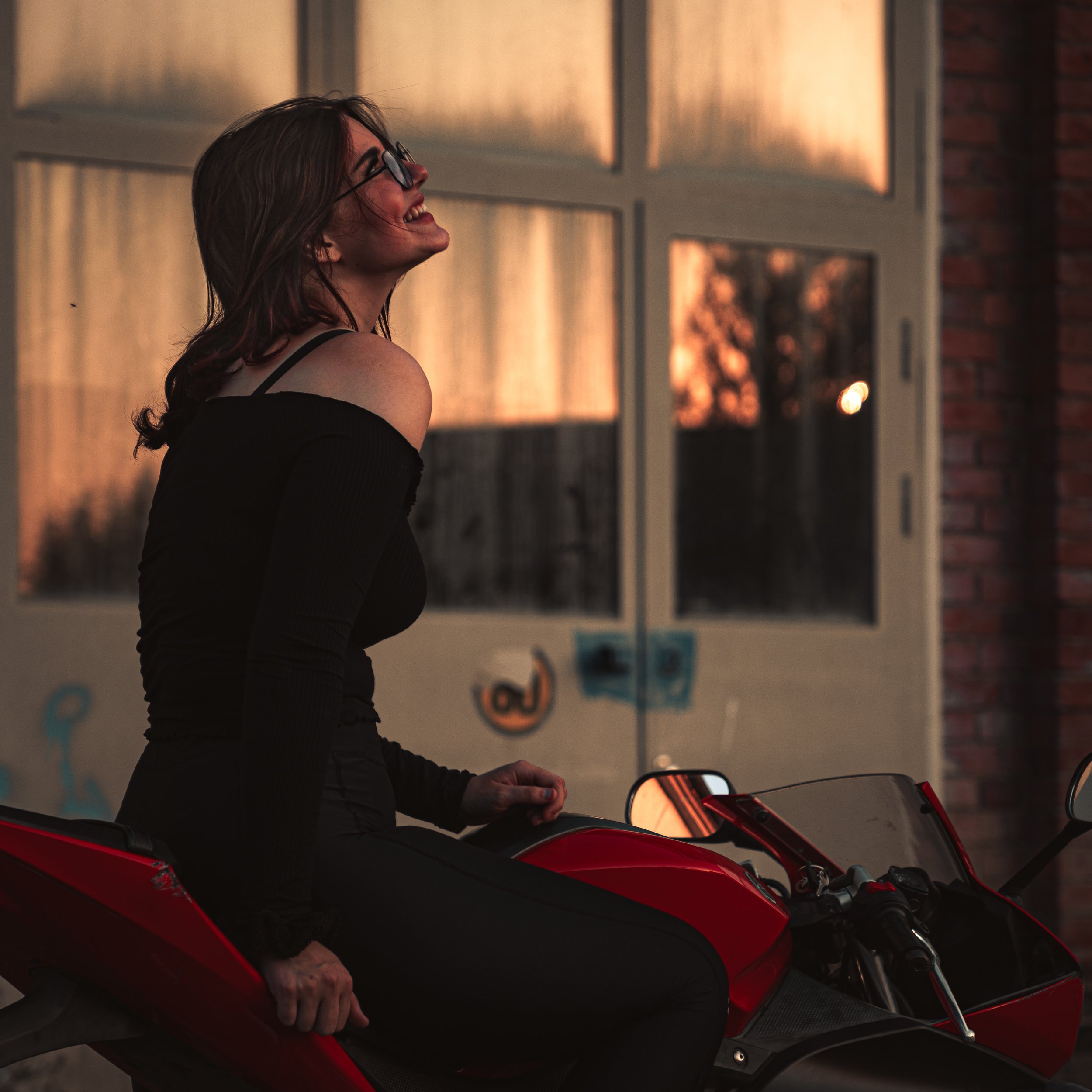 A woman with glasses and brown hair sitting on a red motorcycle outdoors during sunset, smiling and looking up.