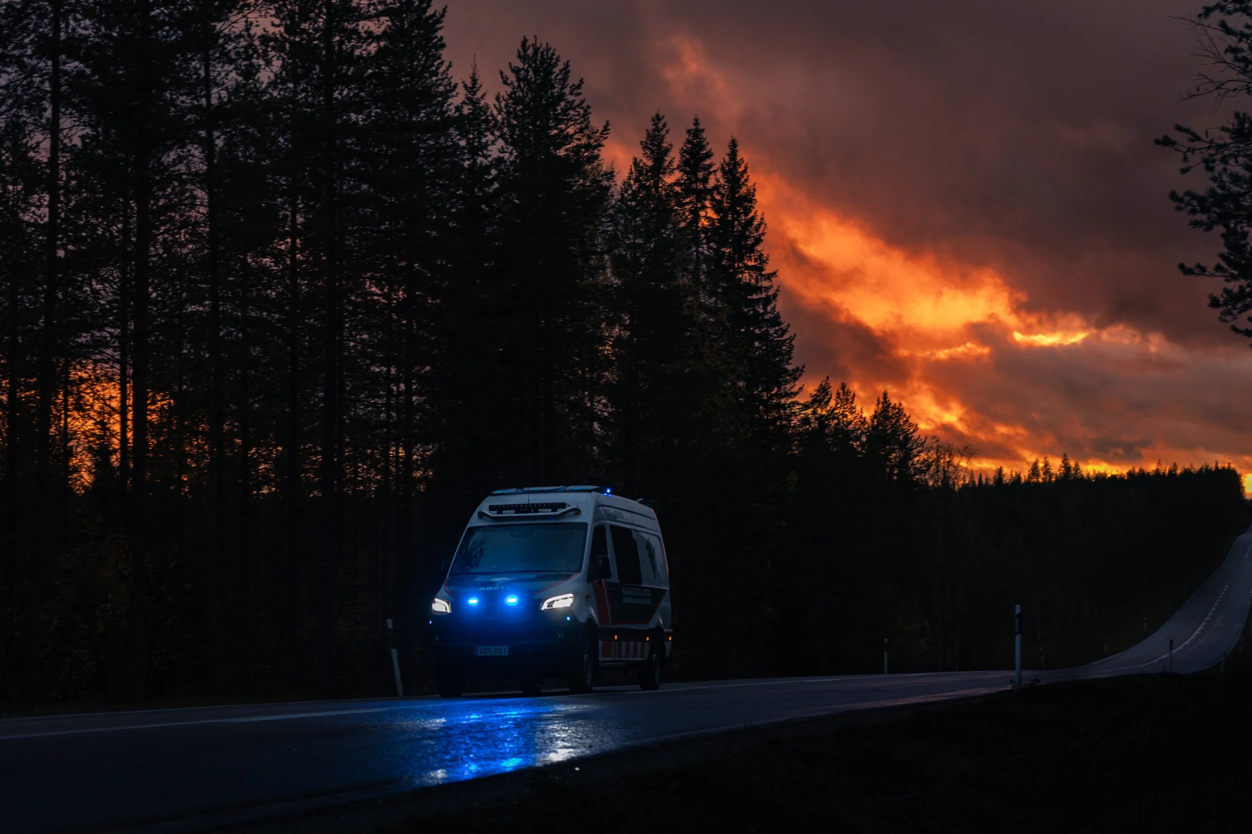 Emergency vehicle on a rural road during sunset with a forest and smoky sky in the background.