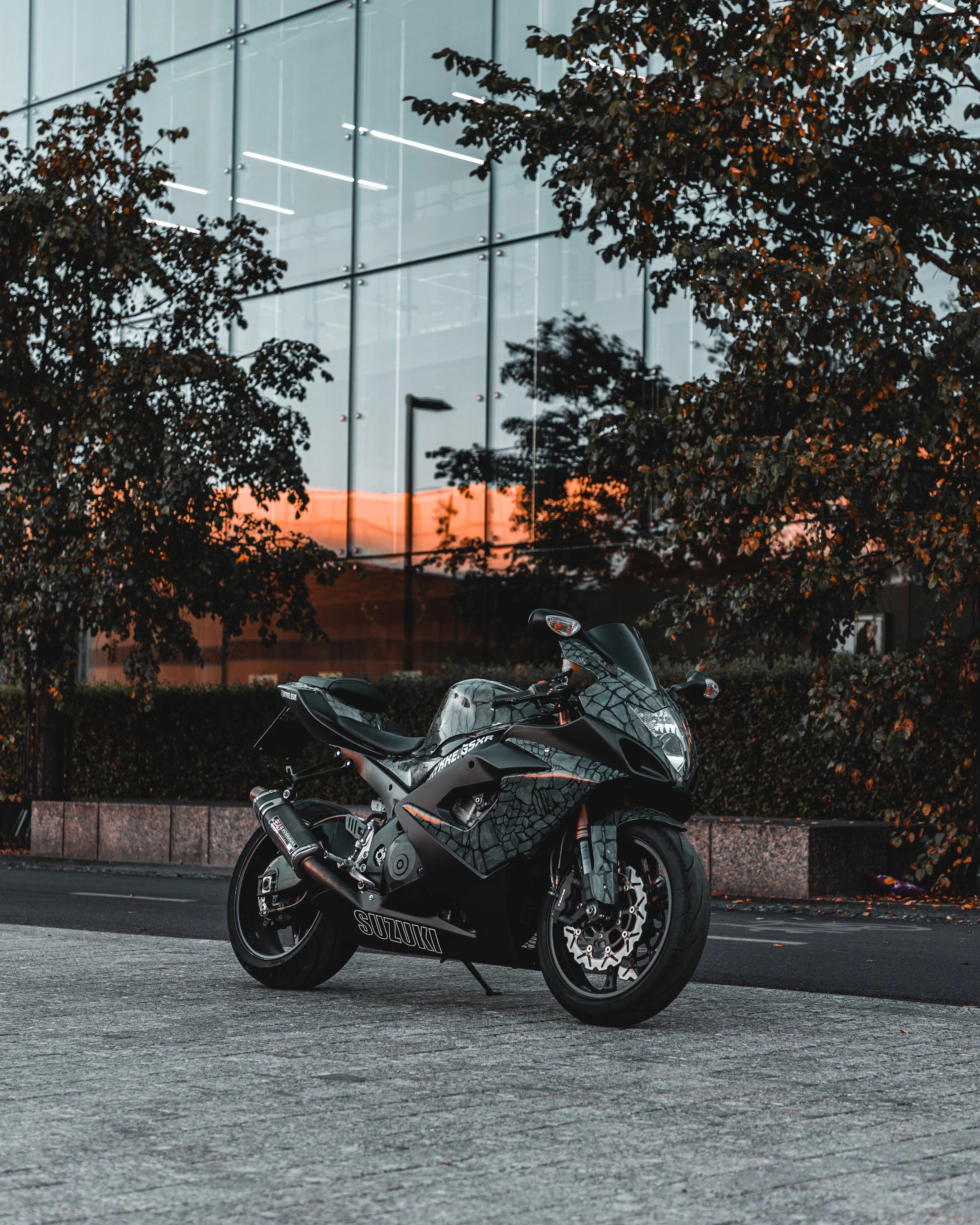 A black Suzuki sport motorcycle parked on a paved area in front of trees and a reflective glass building at sunset.