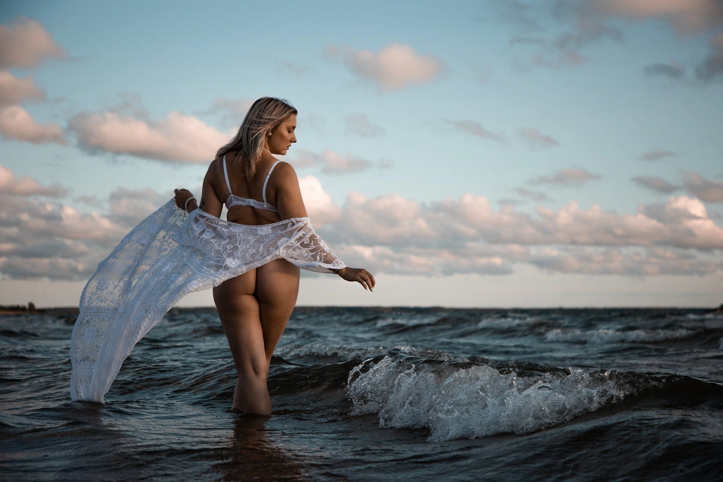 A woman standing in the ocean at sunset, wearing a white lace cover-up and bikini, with her back turned and her hair blowing in the wind.