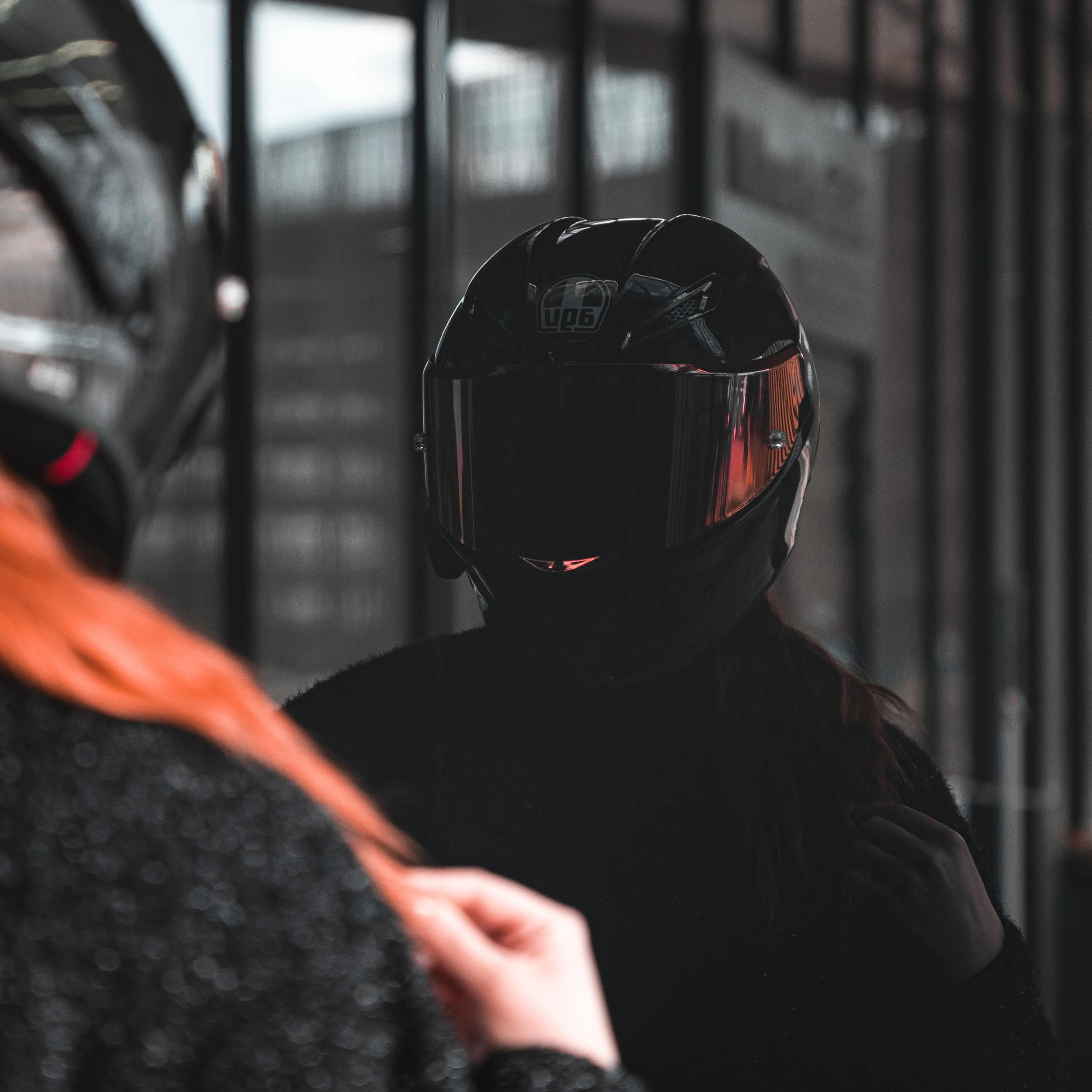 Person wearing a black motorcycle helmet talking to another person with curly red hair, in a dimly lit setting with metal railings.