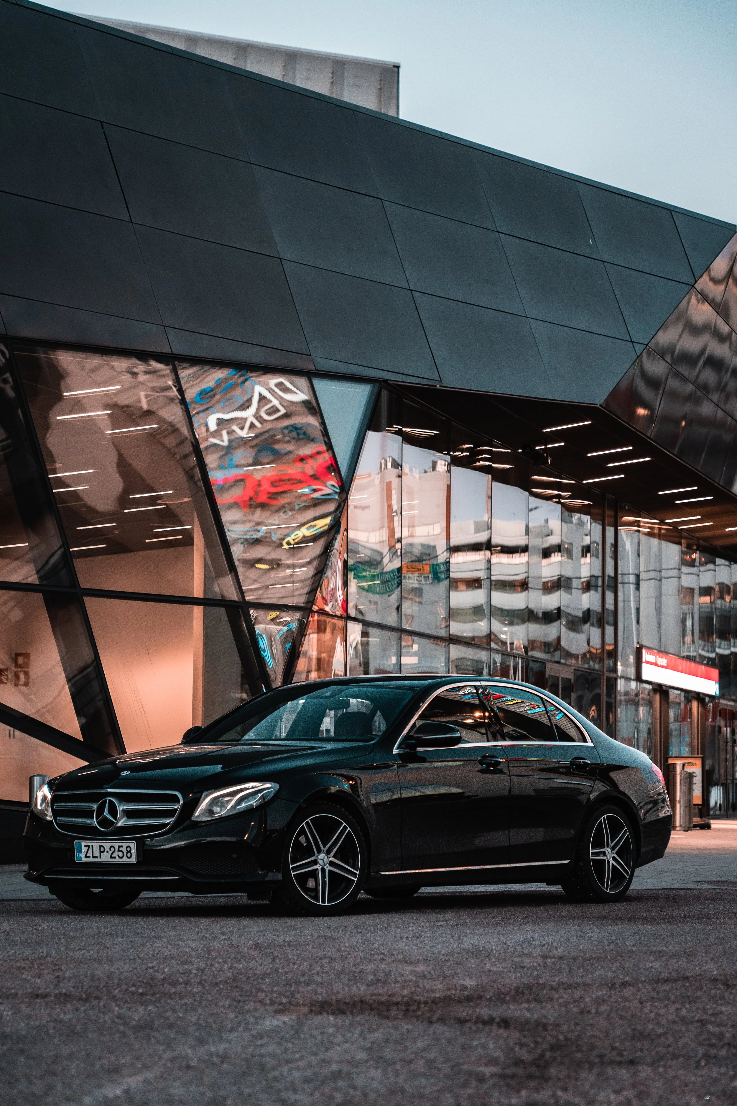 A black Mercedes-Benz car parked outside a modern building with glass walls and reflective surfaces during dusk.