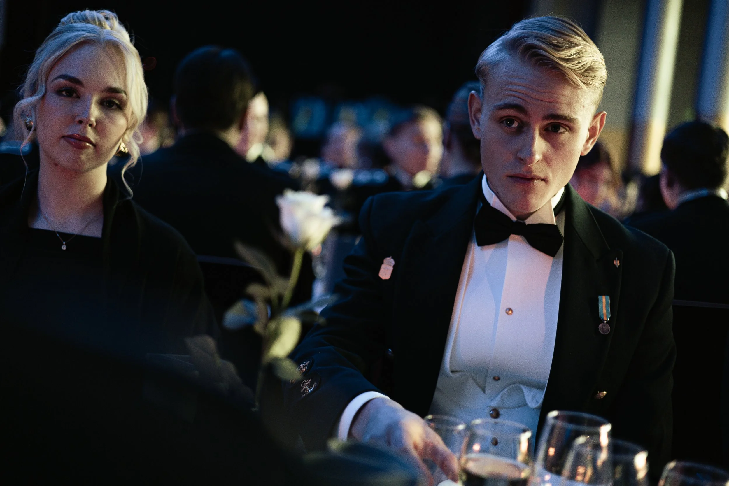 A young man and woman dressed in formal attire, seated at a dinner event with glasses of wine, with a blurred background of other guests.