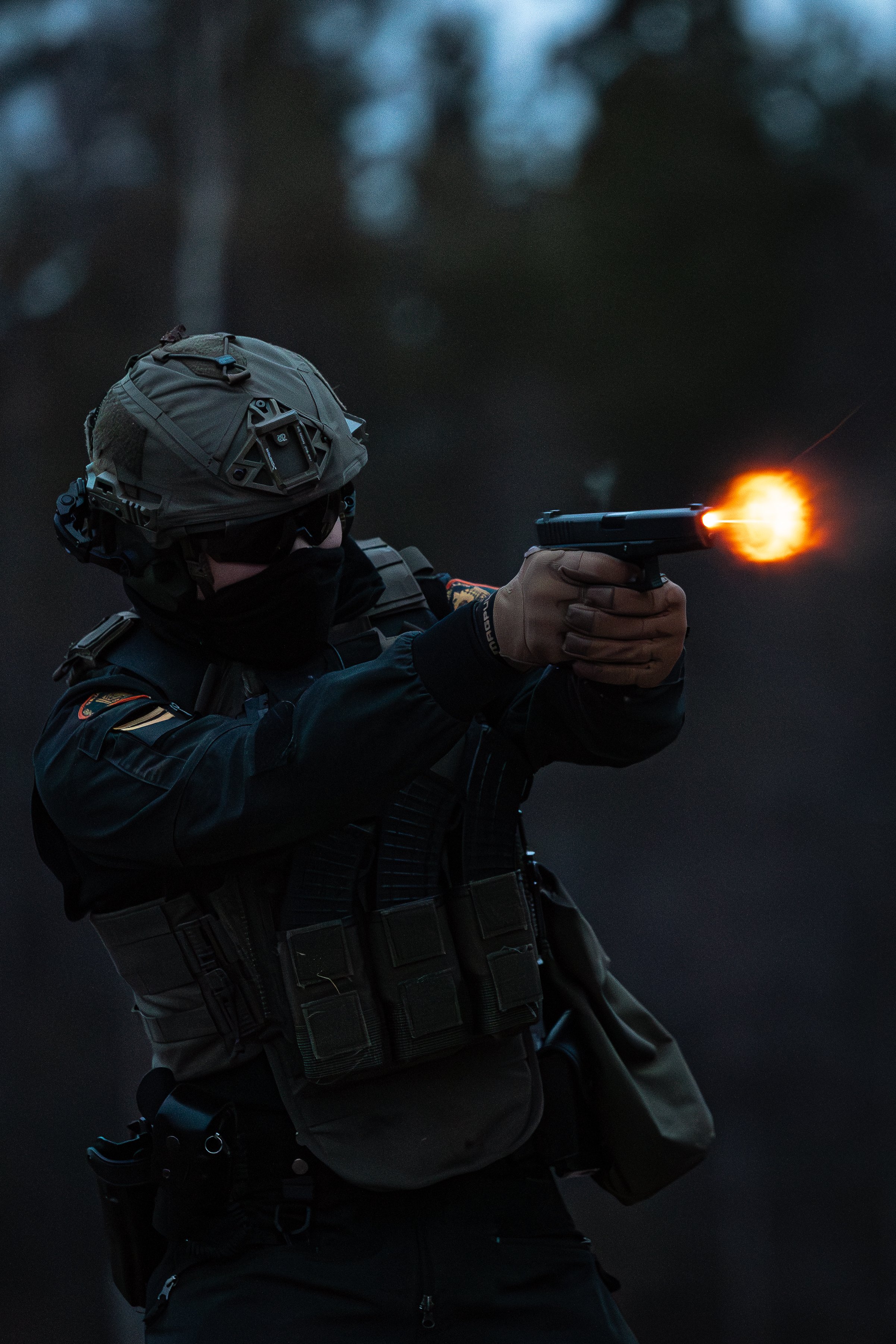 A person dressed in tactical gear shooting a handgun with a visible muzzle flash during dusk or night, outdoors.