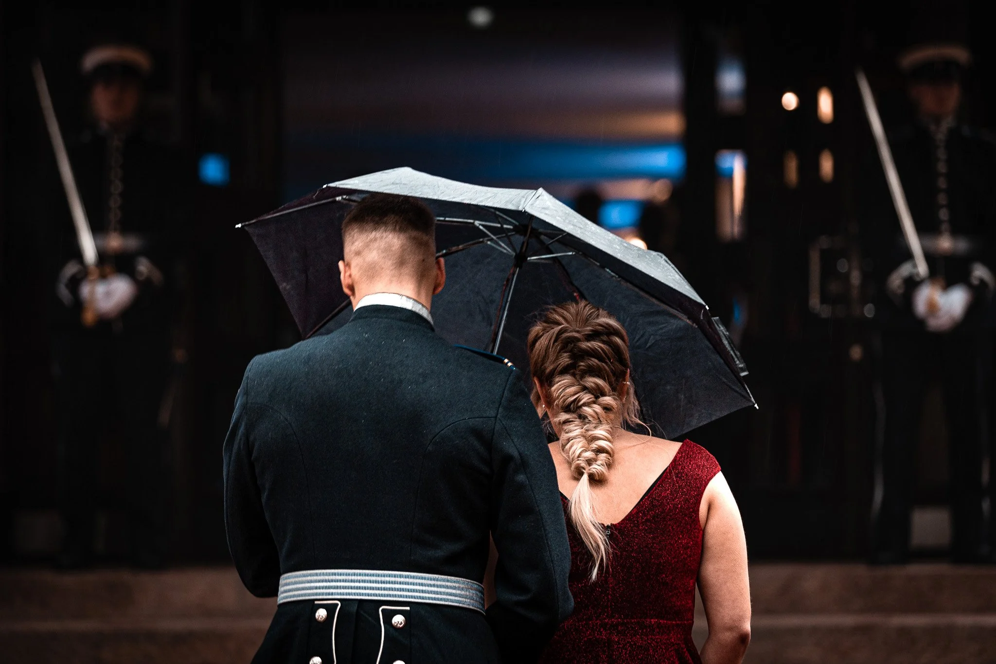 A couple under a black umbrella during a formal event, with military guards in uniform standing in the background.