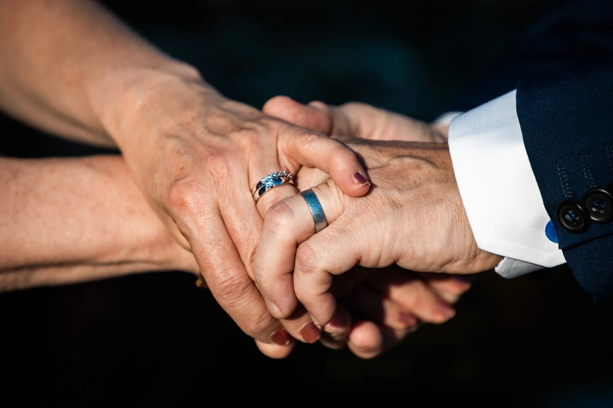 Close-up of a couple holding hands, both wearing wedding rings, one in a suit and the other with rings, symbolizing marriage.