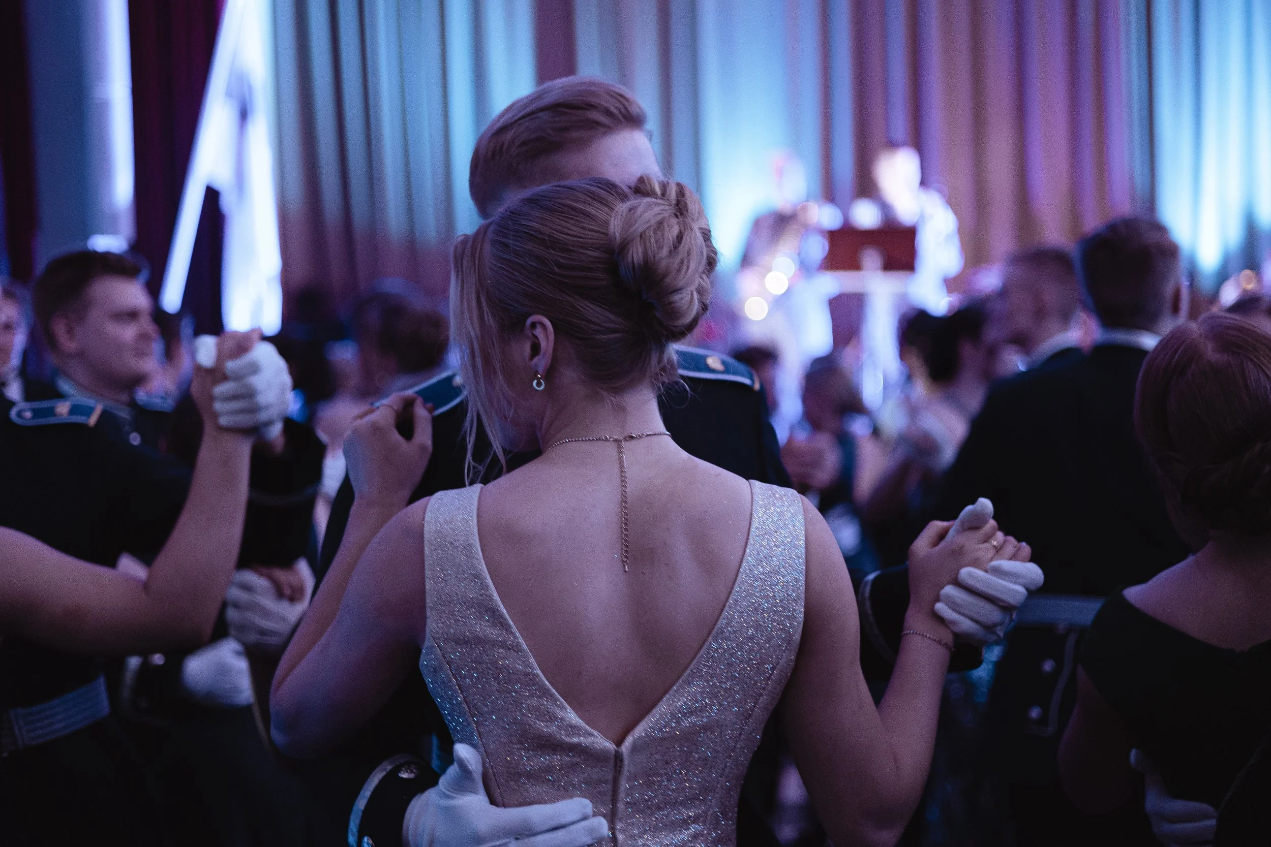 Couple dancing at a formal event, surrounded by other couples in elegant attire, with a live band perform to the back in a dimly lit room with curtains.