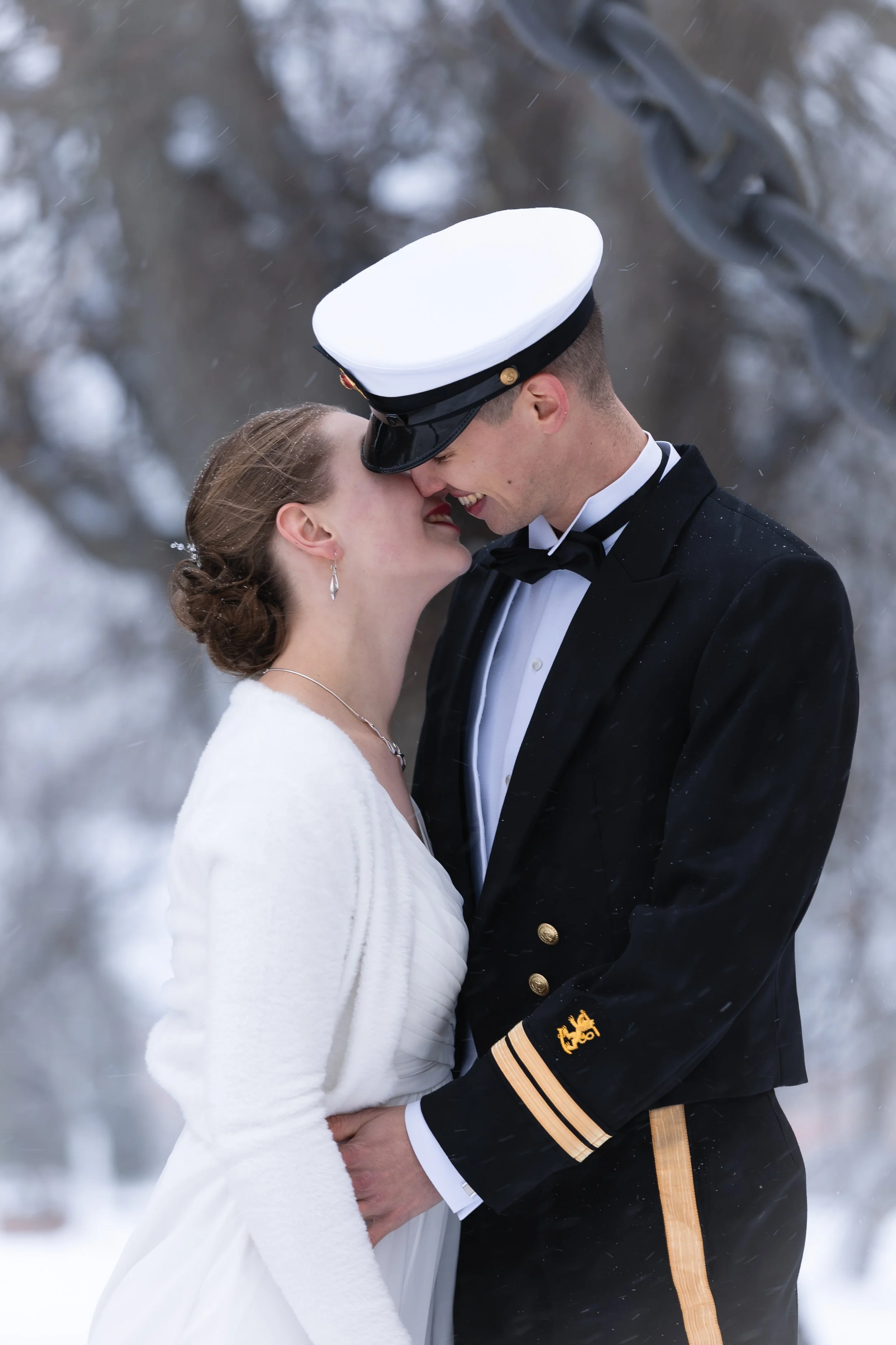 A bride and groom sharing an intimate moment outdoors in winter, with snow and trees in the background. The groom wears a navy captain uniform, and the bride wears a white dress and cardigan.