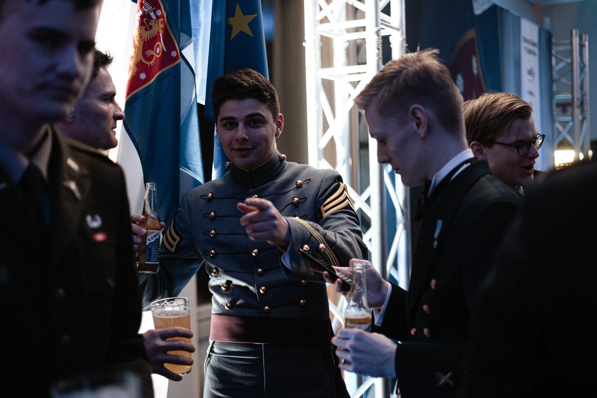 Group of young men in military uniforms at a formal event, with flags in the background, holding drinks and looking at a phone.