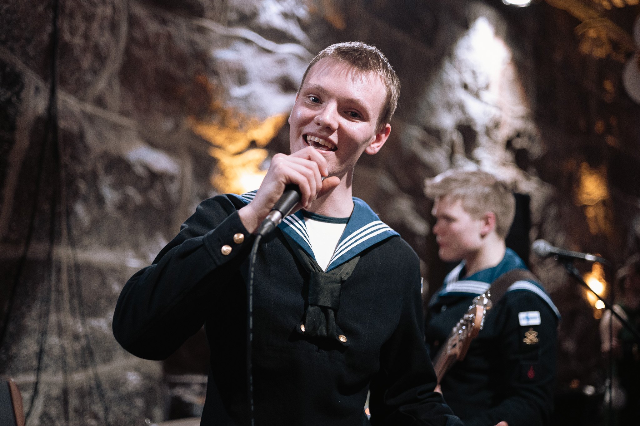 A young man in a navy sailor uniform holding a microphone and smiling, with other musicians in similar uniforms playing instruments in the background.