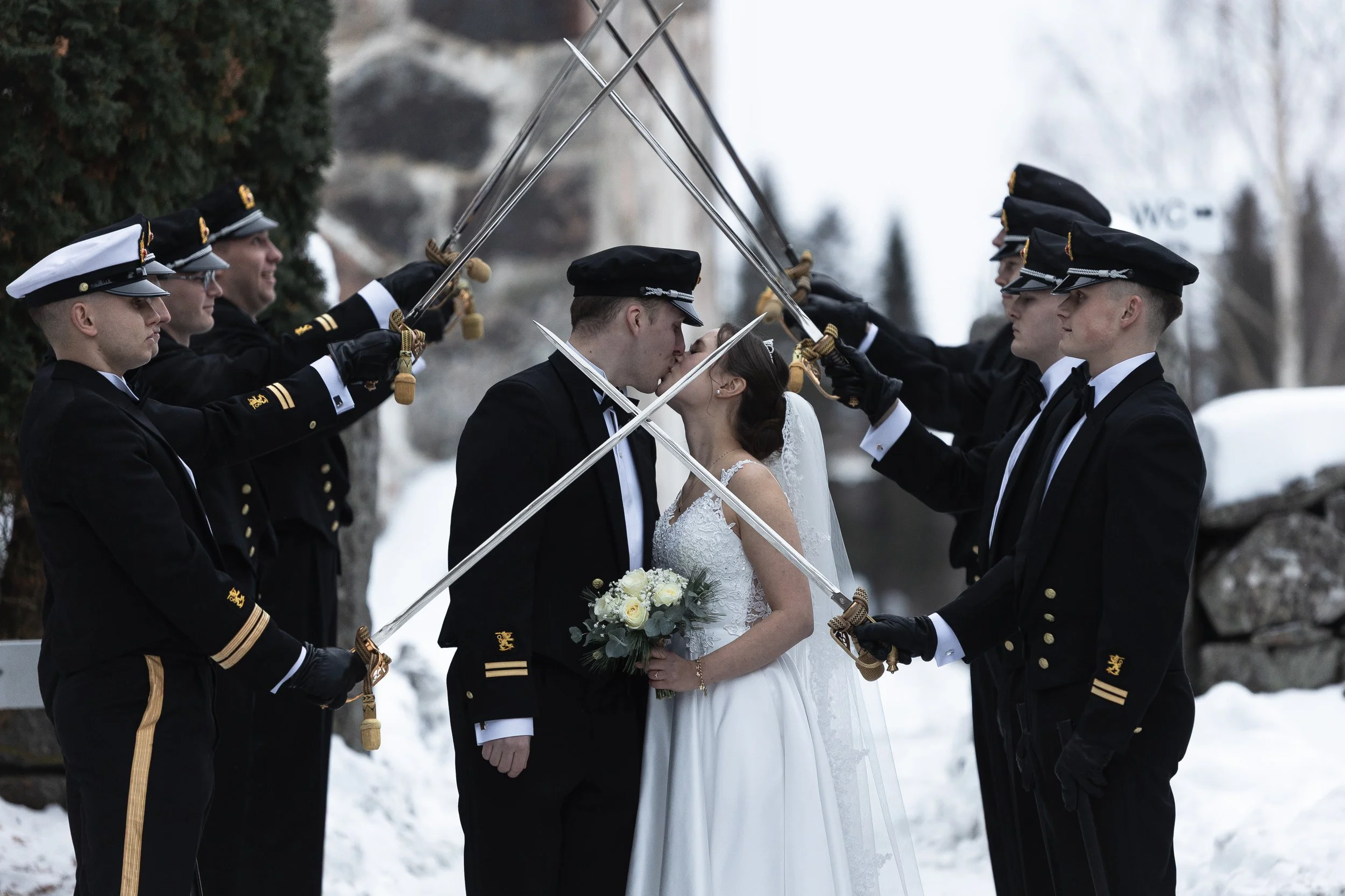 Bride and groom kissing under swords held by naval officers in uniform outside in snow.
