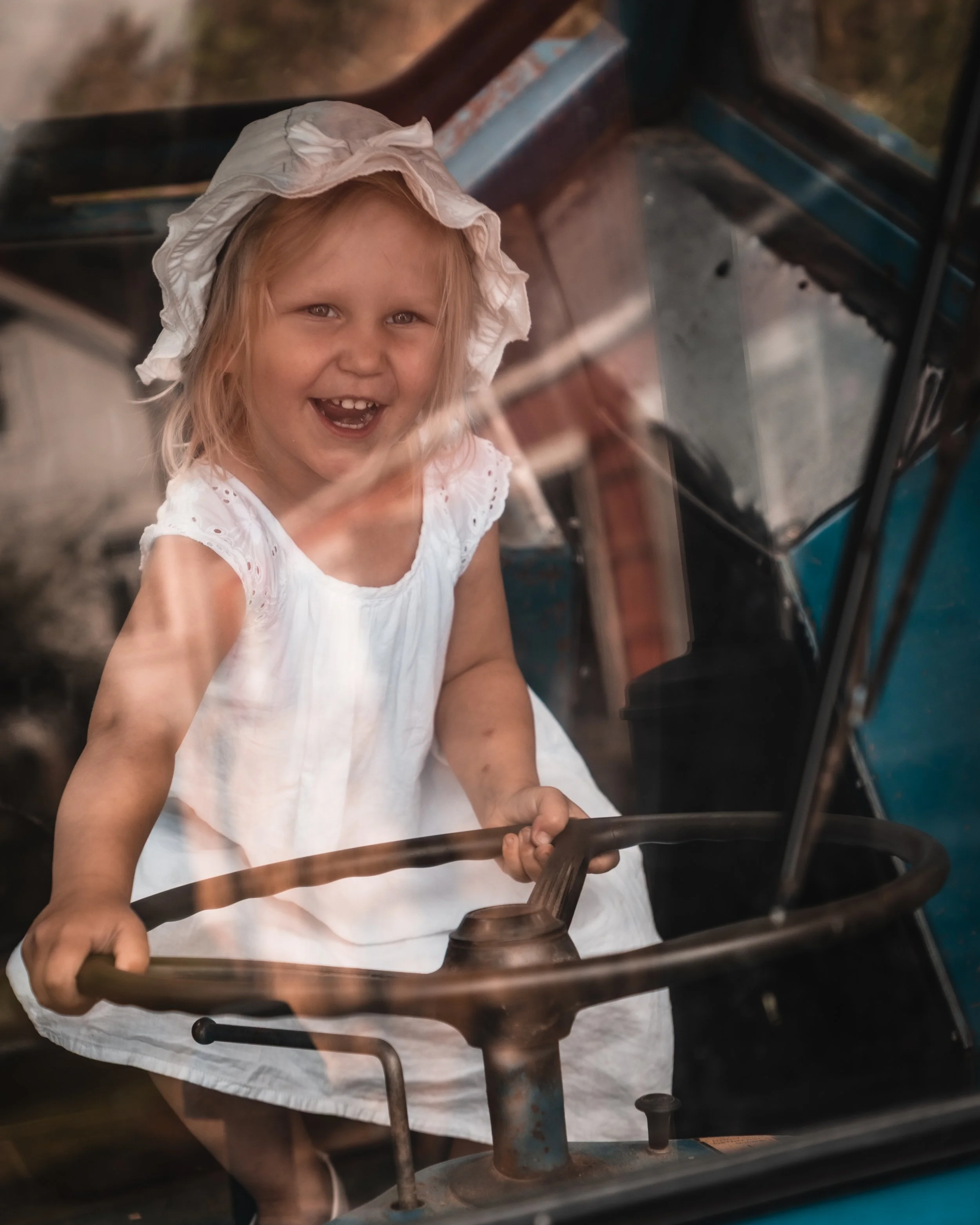 A young girl with blonde hair wearing a white dress and a white bonnet, smiling and sitting in the driver's seat of an old vehicle, holding the steering wheel.