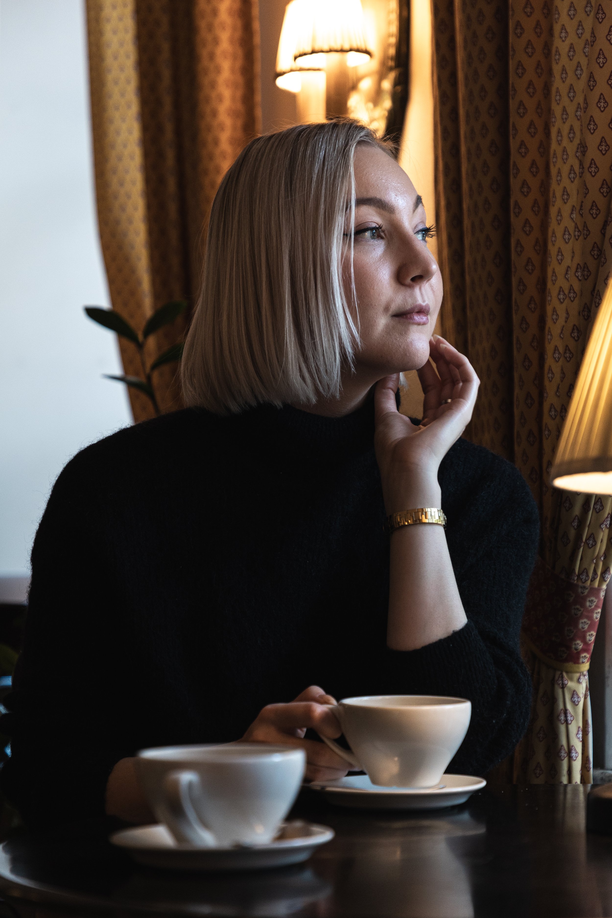 A woman with shoulder-length blonde hair sitting at a table in a cozy cafe, holding a teacup and looking thoughtfully to the side near a window with patterned curtains and warm lighting.