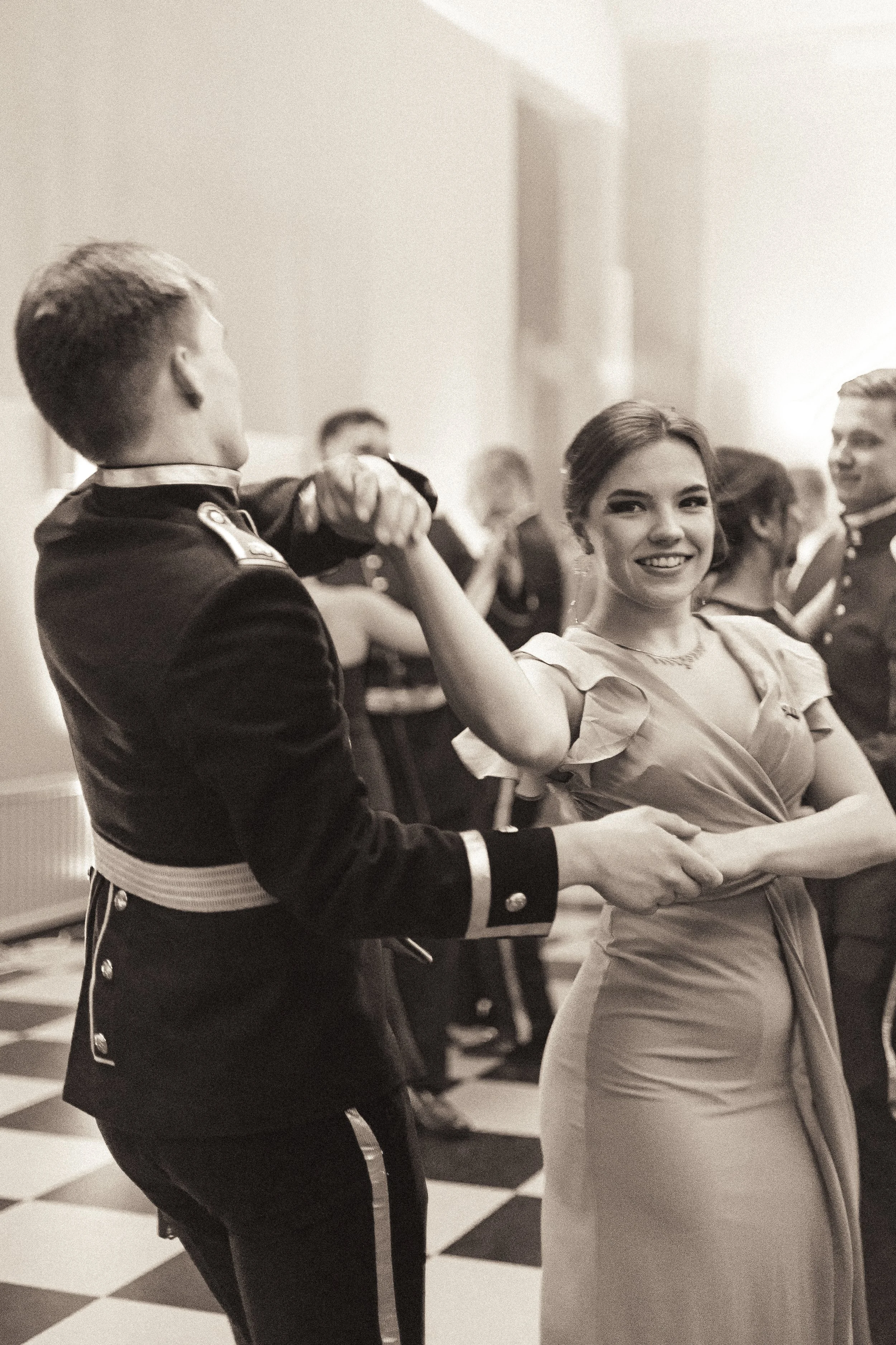 A black and white photo of a couple dancing at a formal event, with other couples dancing in the background.
