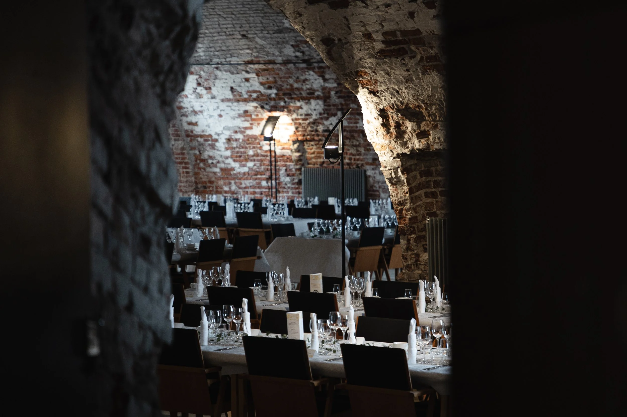 Dinner party setup in a rustic brick cellar with long tables, glassware, and napkins, illuminated by standing lamps.