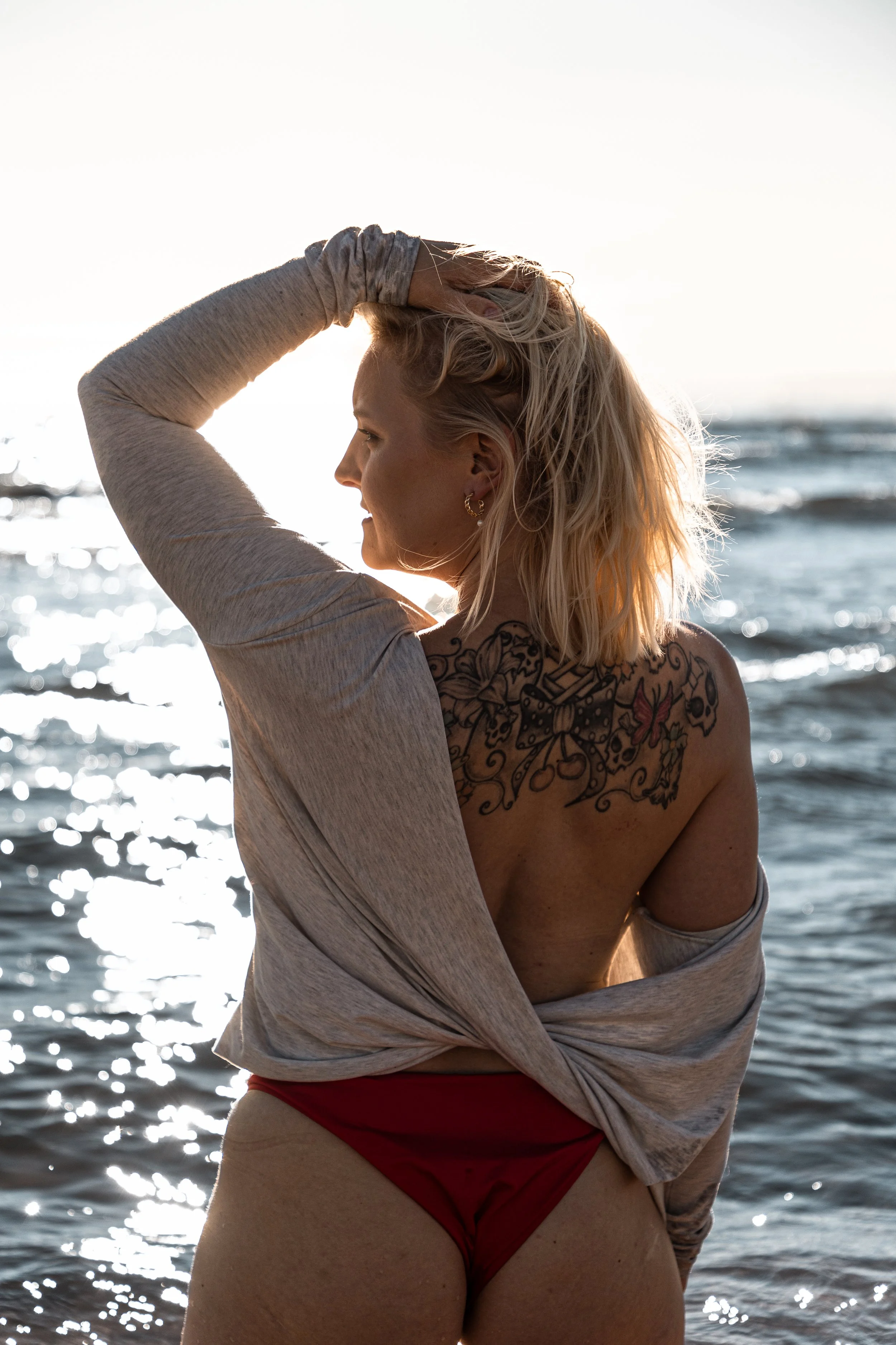 A woman with blonde hair and tattoos on her shoulder standing by the ocean at sunset.