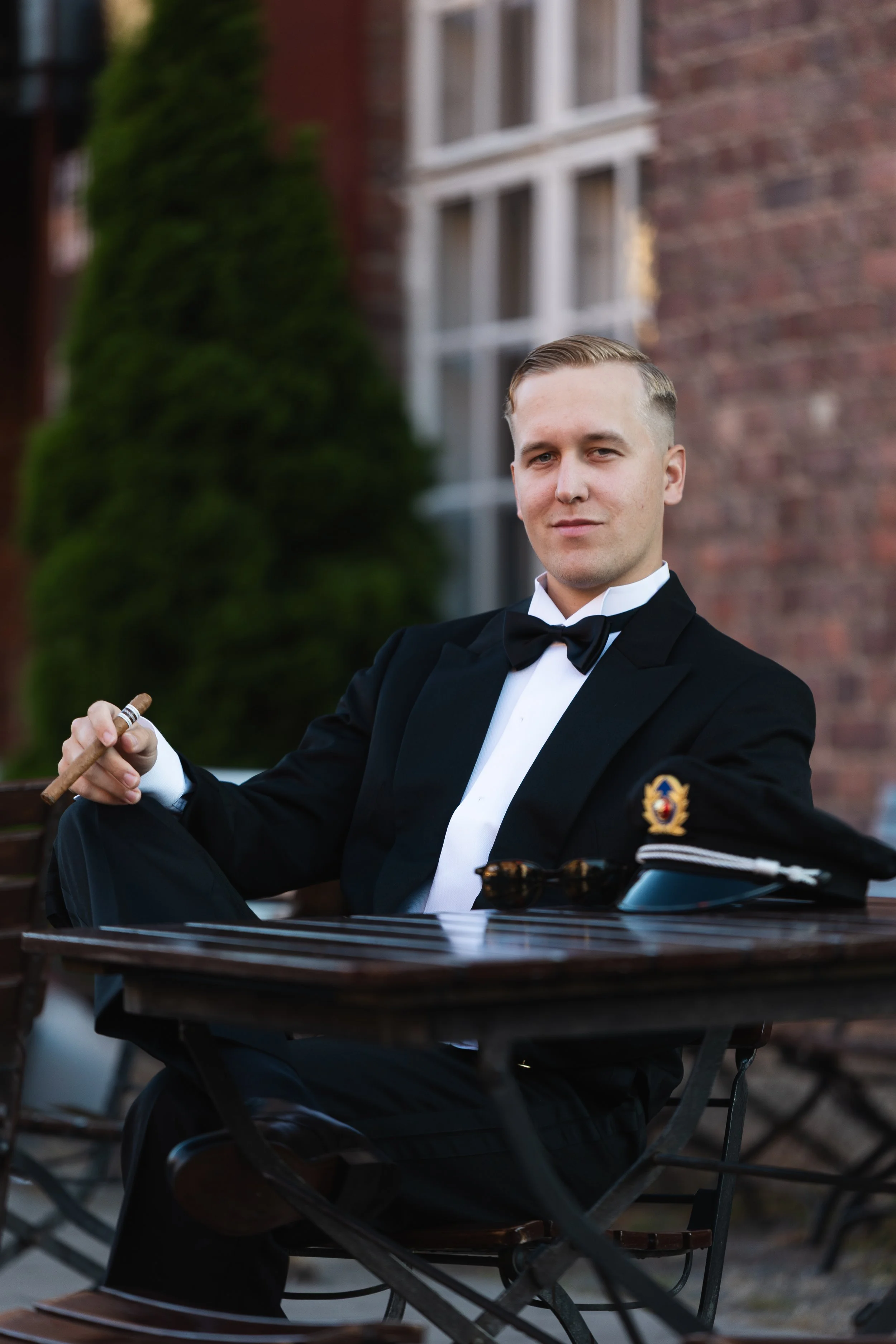 A young man in a tuxedo with a badge on his sleeve, holding a cigar, sitting outdoors at a table with a brick building and greenery in the background.